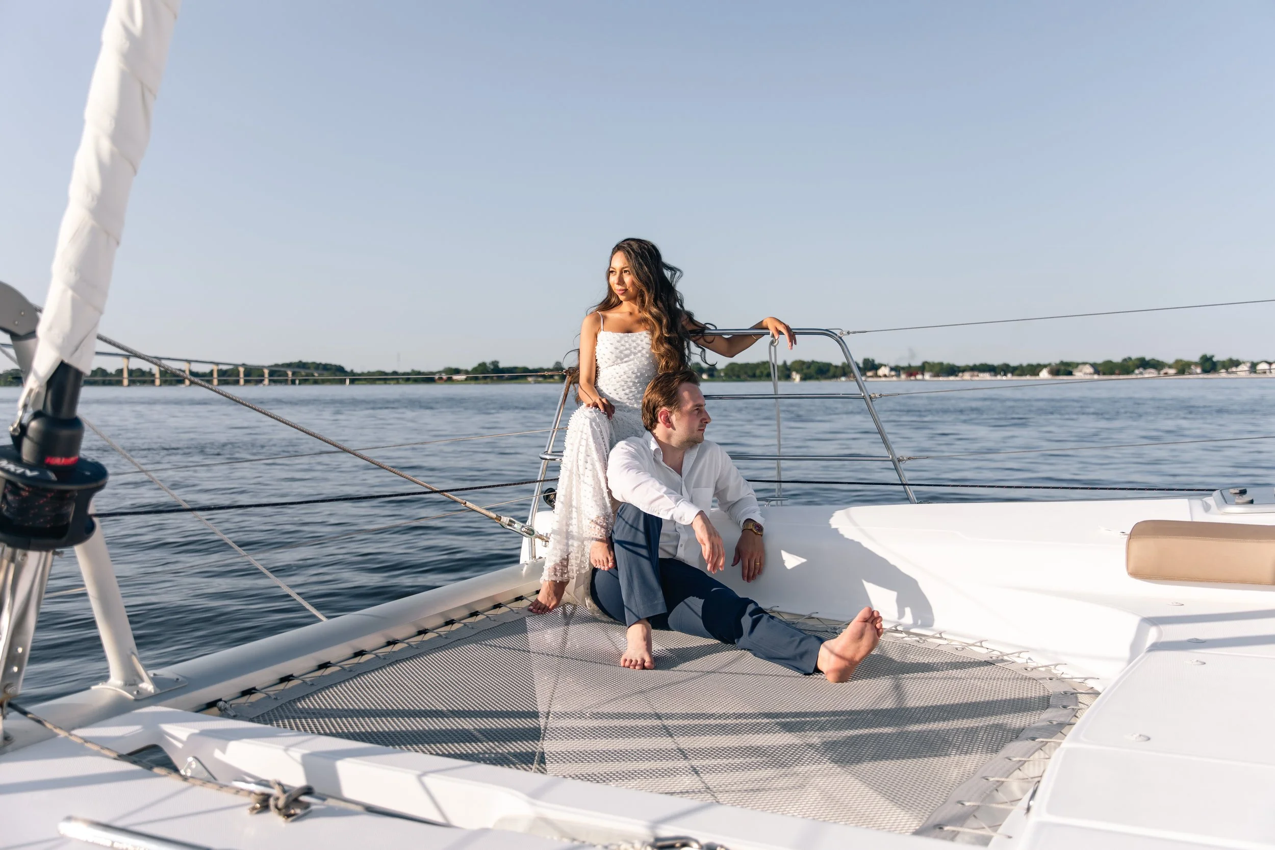 A couple on a sailboat, with the woman standing and the man sitting on the trampoline of the boat, enjoying a day on the water.
