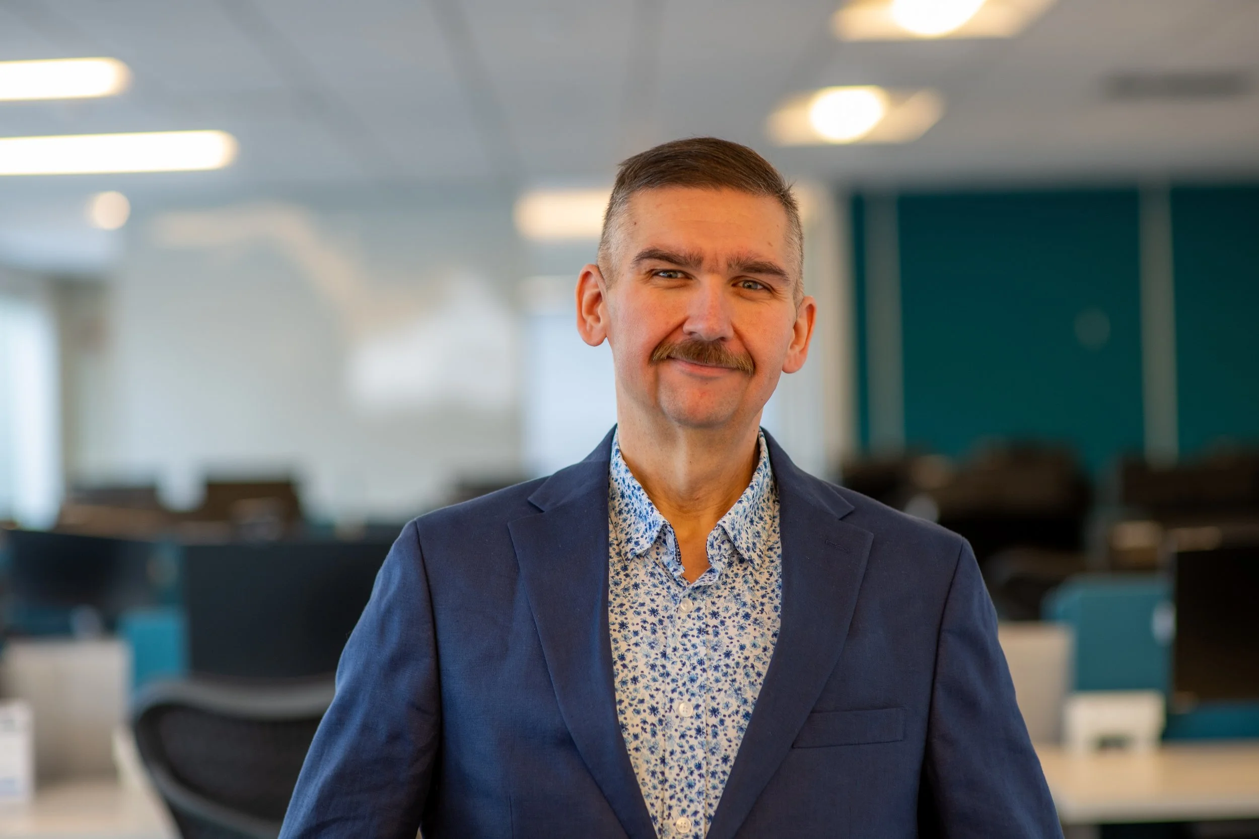 A man with a mustache in a blue blazer and a patterned shirt standing in an office, smiling at the camera.
