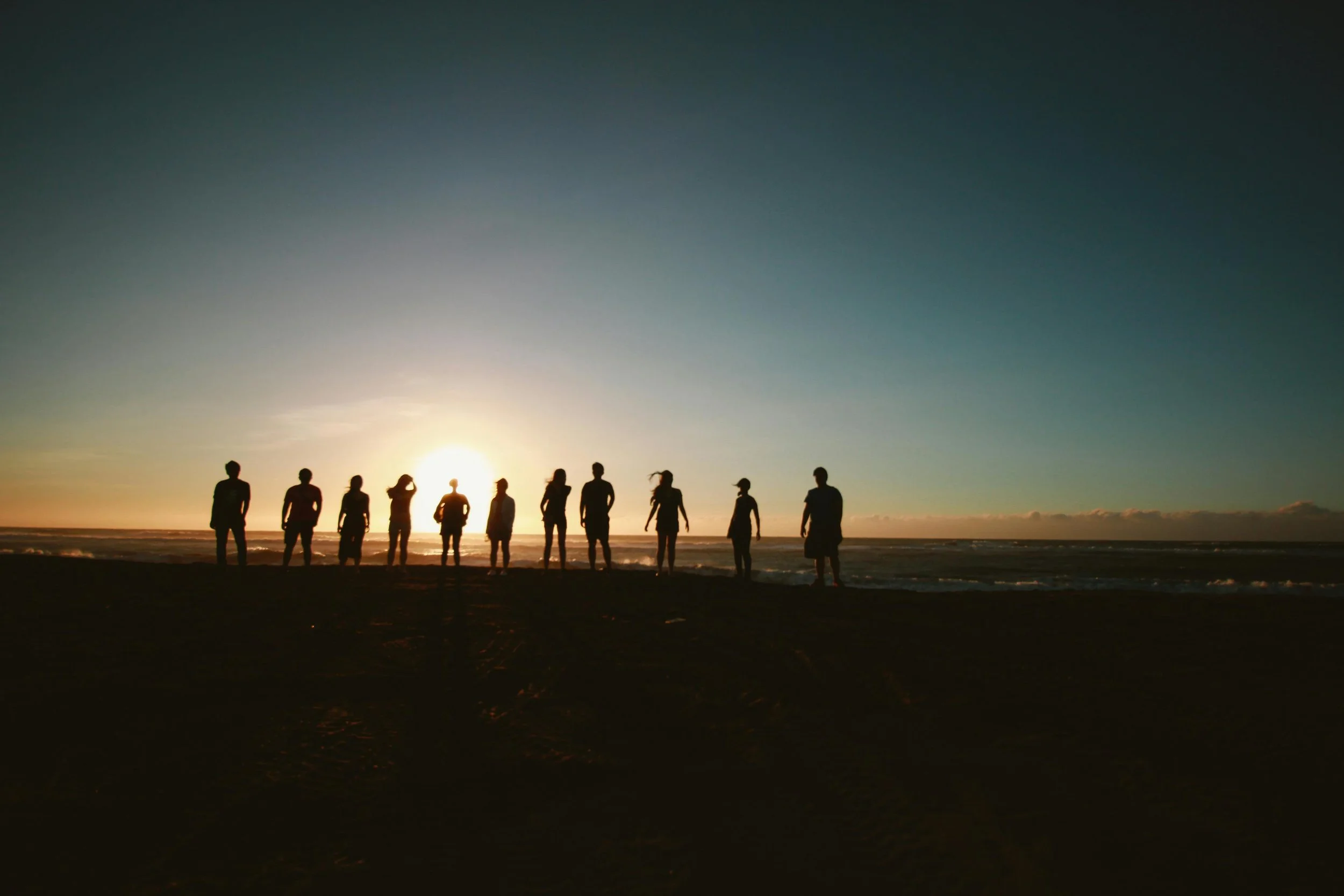 Silhouettes of ten people standing on a beach at sunset, facing the ocean.