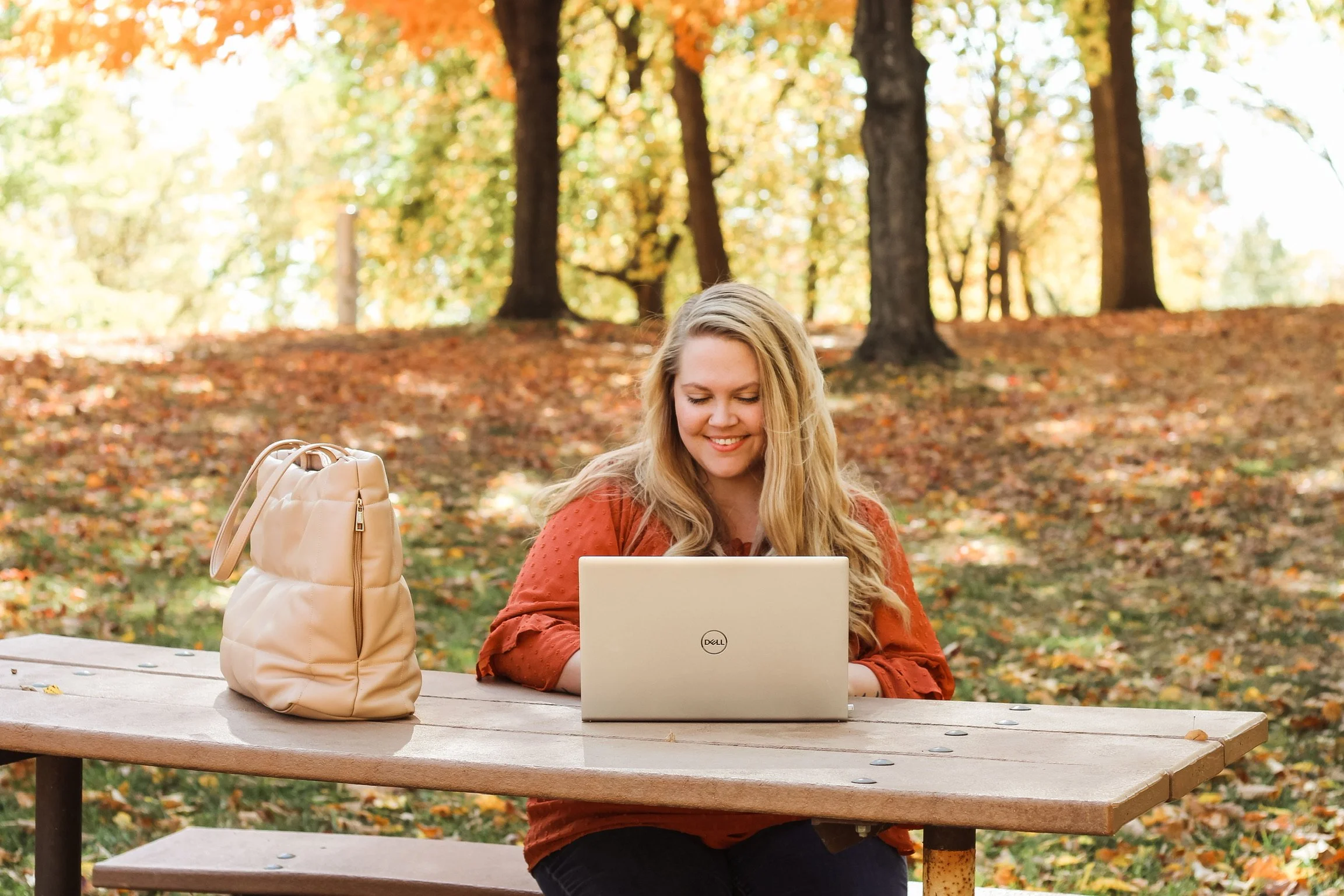 A woman with long blonde hair sitting at a wooden picnic table outdoors, working on a silver laptop, with a beige backpack beside her, in a park with trees and autumn leaves.
