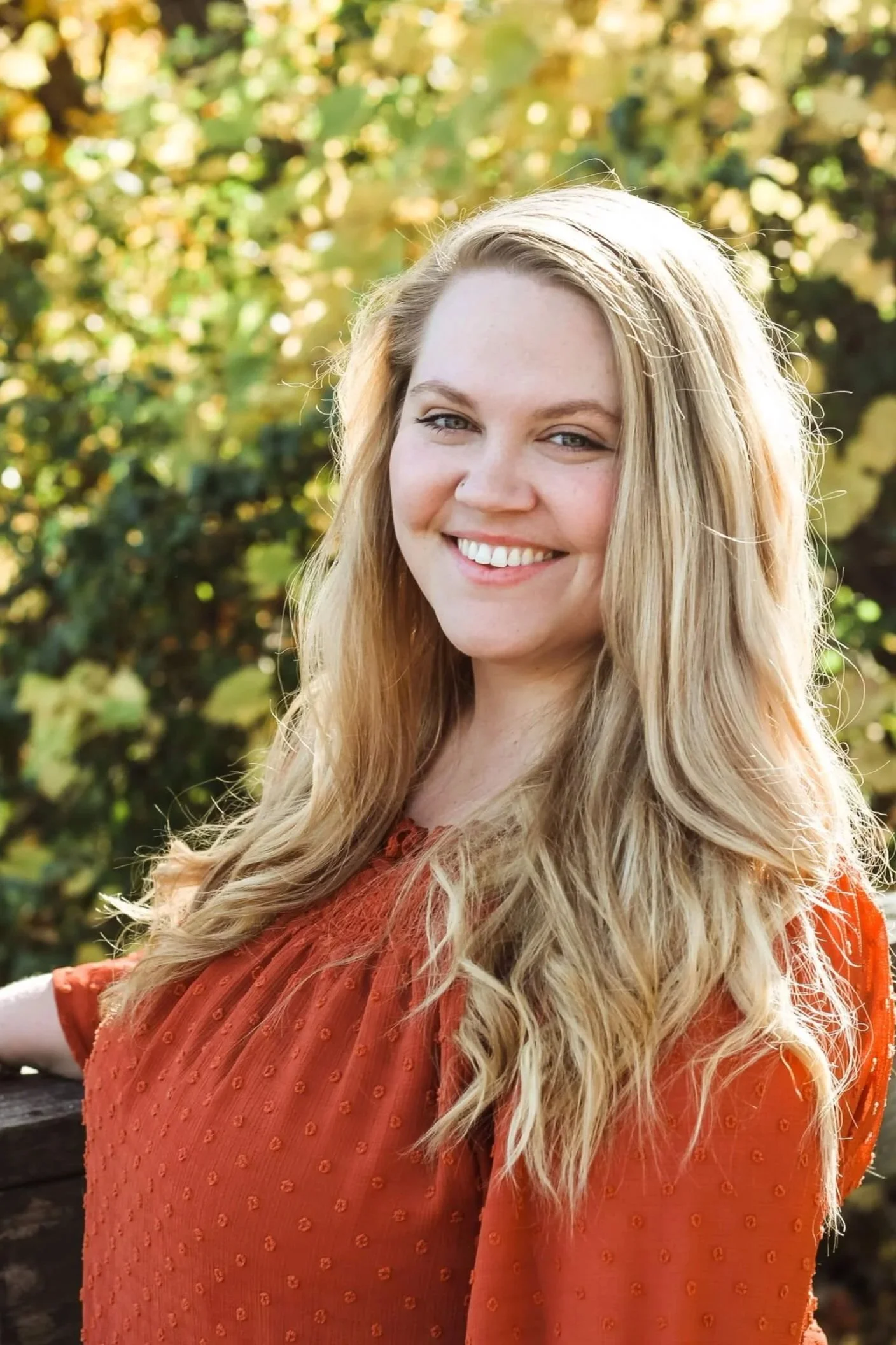 A smiling blonde woman with long wavy hair, wearing an orange top, standing outdoors with fall-colored foliage in the background.