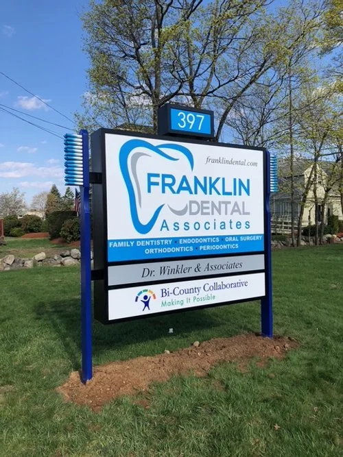 Sign for Franklin Dental Associates with a tooth graphic and listing of dental services, located near trees and a residential area.