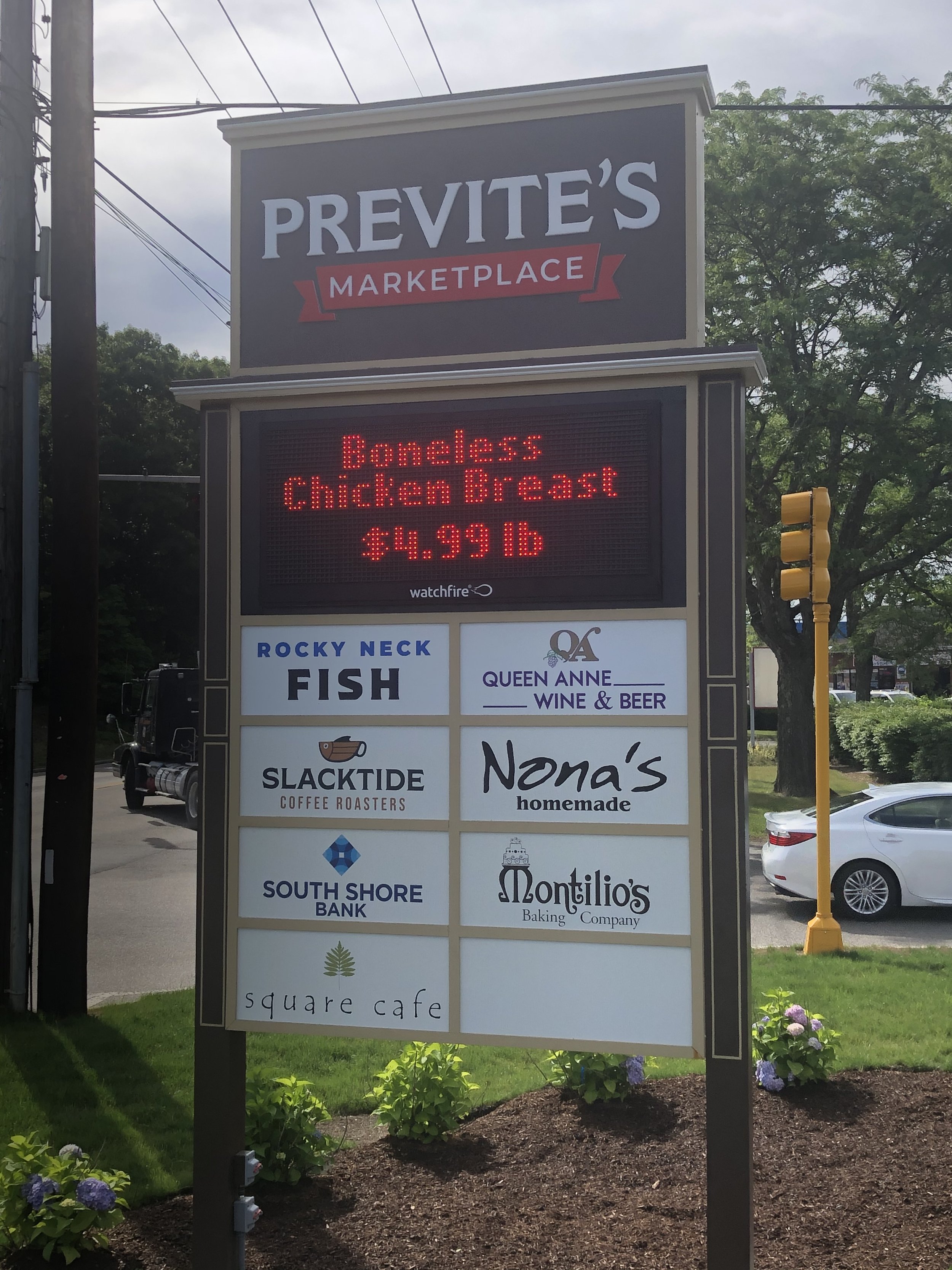 Signboard for Previte's Marketplace, with advertisements for local businesses including Rocky Neck Fish, Queen Anne Wine & Beer, Slackside Coffee Roasters, Nana's Homemade, South Shore Bank, Montilio's Baking Company, and Square Cafe, with trees, car