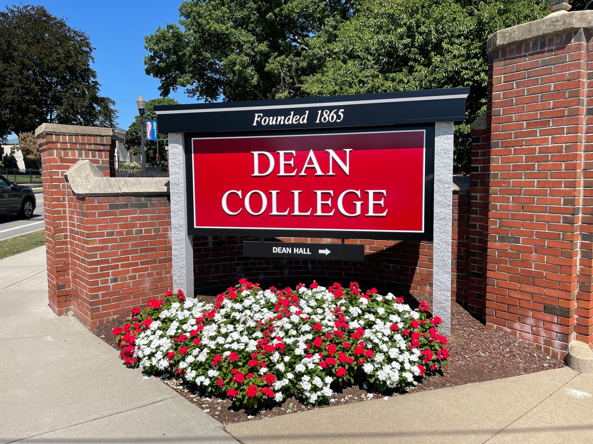Dean College custom monument sign with brick pillars, red and white flowers, and a black and red background, with text indicating the college was founded in 1865 and directing to Dean Hall.