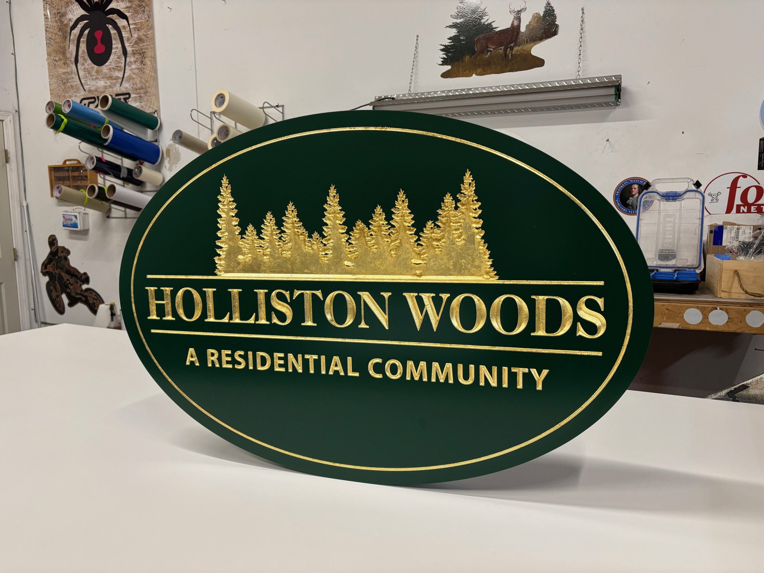 A green oval sign with gold lettering and gold trees reads "HOLLISTON WOODS A RESIDENTIAL COMMUNITY" in an indoor workshop setting.