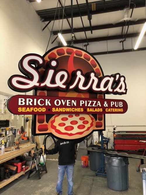 Large sign for Sierra's Brick Oven Pizza & Pub, featuring a pizza image and text advertising seafood, sandwiches, salads, and catering, inside a commercial kitchen or workshop.
