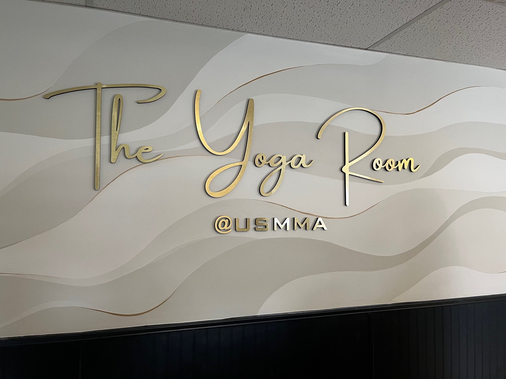 Sign on a wall reading 'The Yoga Room' with the Instagram handle beneath, in a stylish gold font against a textured white background.
