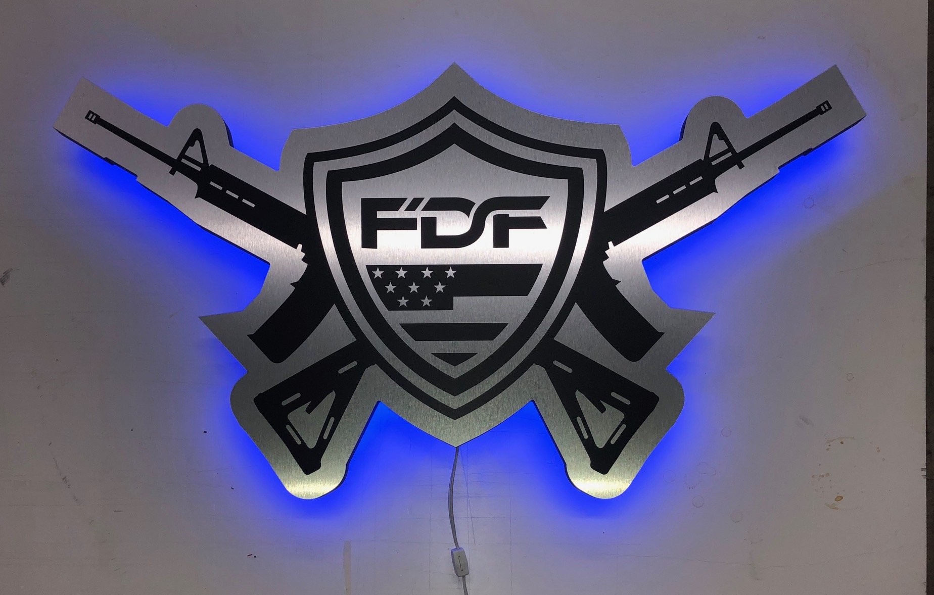 A wall-mounted sign featuring crossed rifles, a shield with a flag, and the letters FDF, illuminated by blue backlighting.