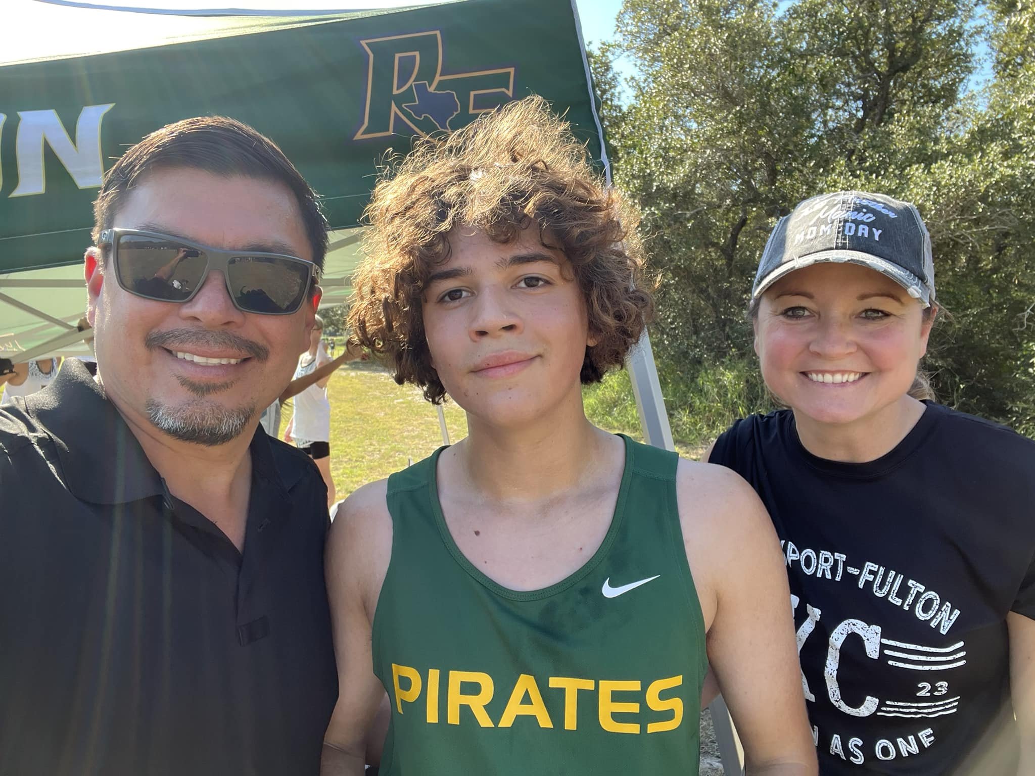 Three people taking a selfie outdoors, one in a green sports jersey with 'Pirates' on it, flanked by a man wearing sunglasses and a woman wearing a cap and a t-shirt, with a green canopy and trees in the background.