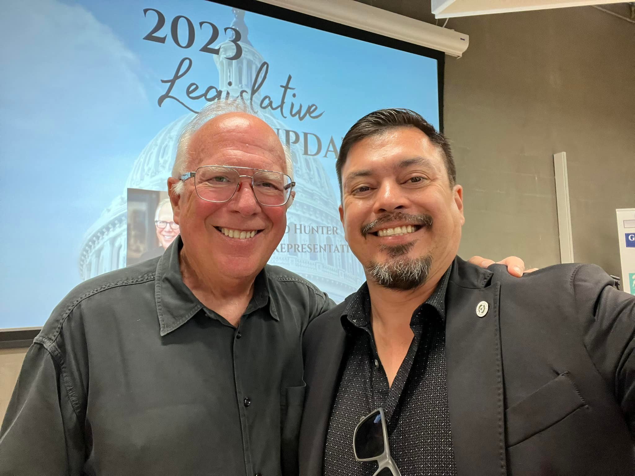Two men taking a selfie at a 2023 legislative update event, standing in front of a presentation screen with a blurred image of the US Capitol.