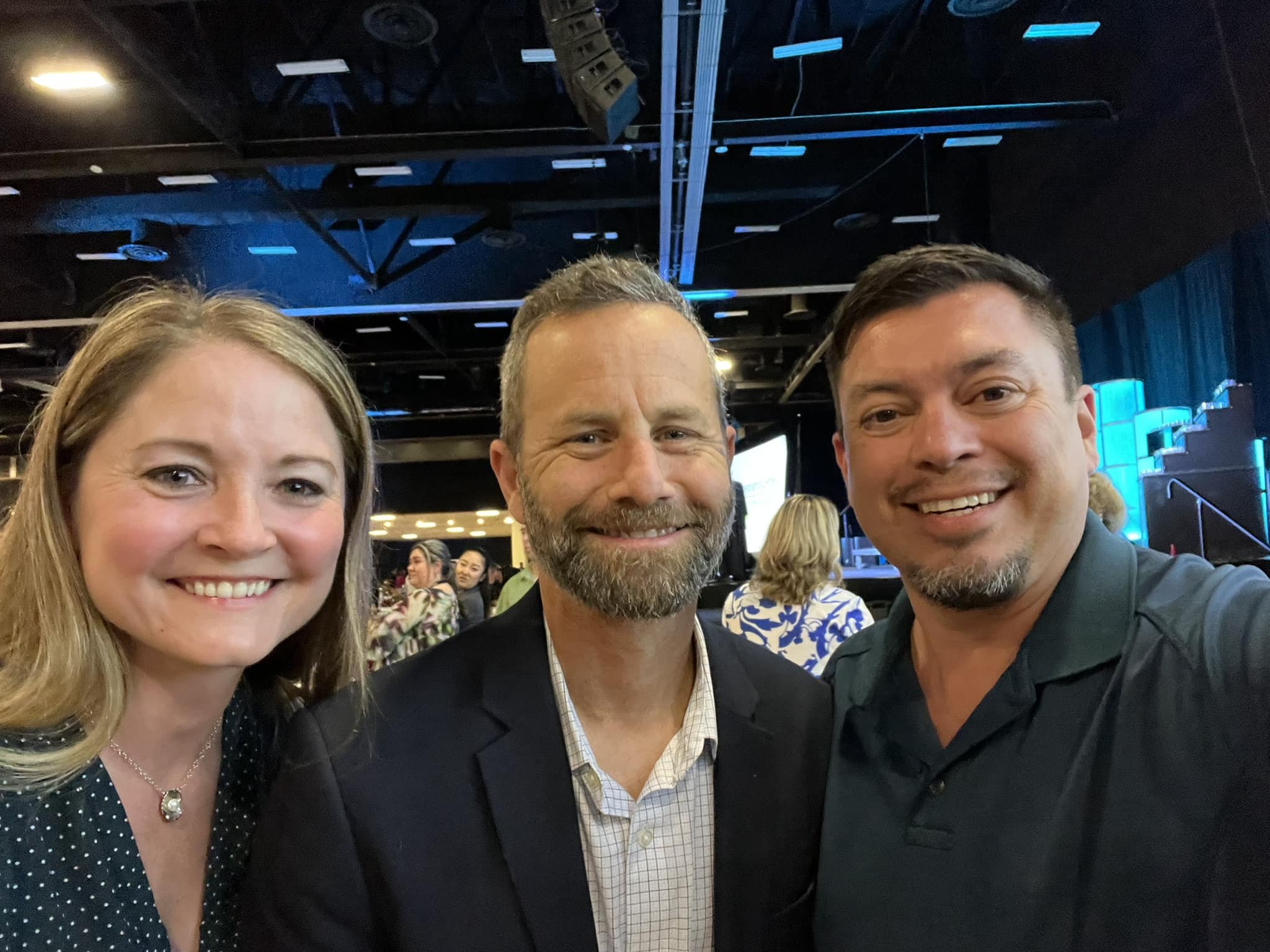 Three people taking a selfie together at an indoor event, smiling with a dark venue background and other people in the distance.