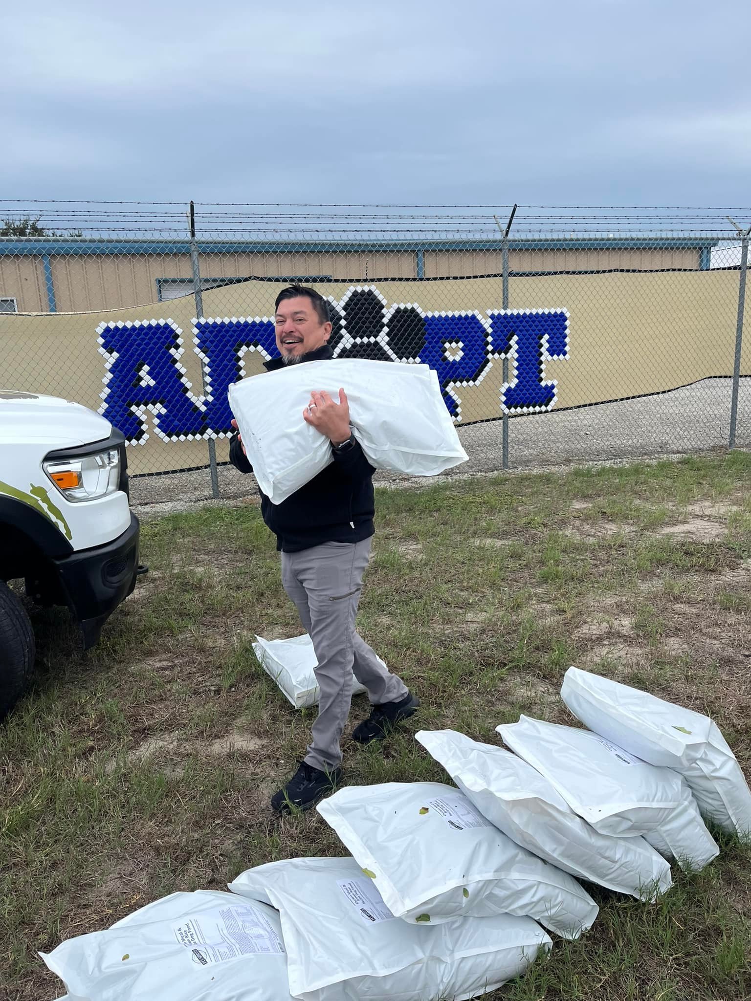 A man holding a white pillow standing on grass in front of a chain-link fence with a blue and black mural spelling 'ALPHA'. Multiple white pillows or comforters are on the ground around him, and a white vehicle with a green emblem is partially visibl