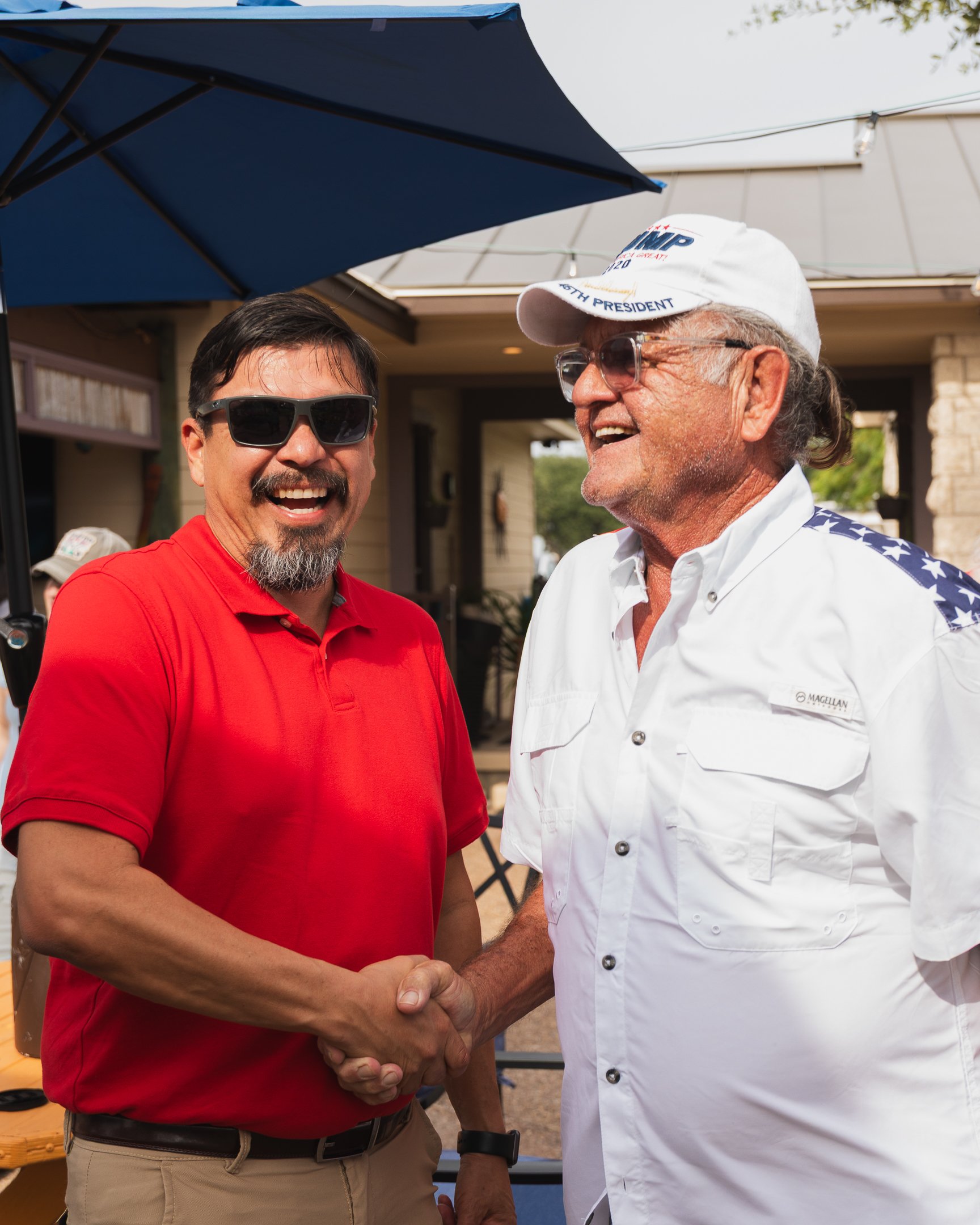 Two men shaking hands outdoors, one wearing a red shirt and sunglasses, and the other wearing a white shirt with stars on the shoulders and a white cap with 'TRUMP' and 'PRESIDENT' written on it, smiling and engaging in friendly conversation.