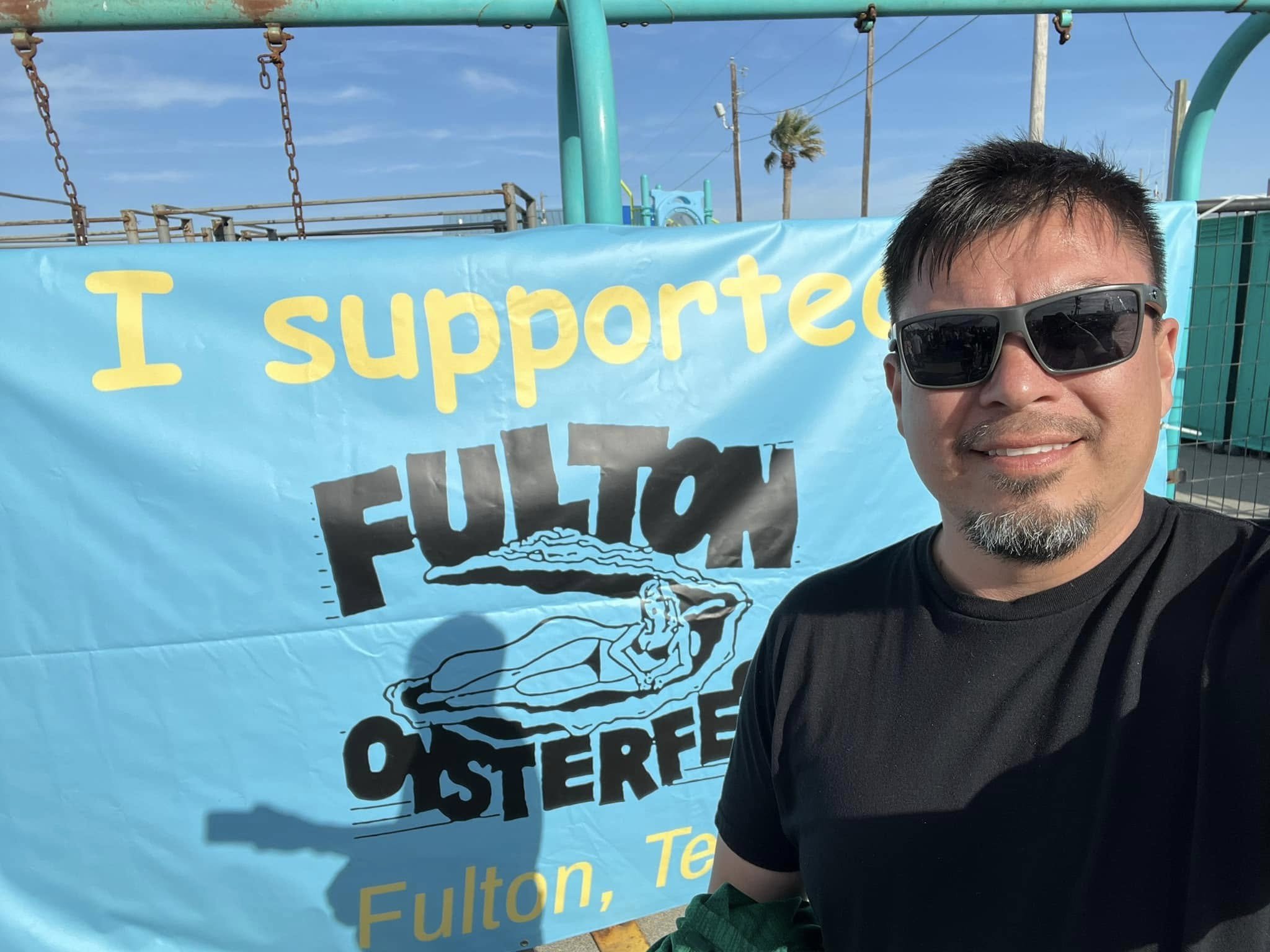 Man in black t-shirt and sunglasses smiling in front of a Fulton Oyster banner outdoors.