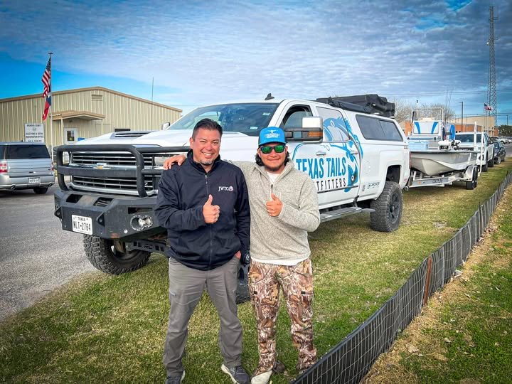 Two men standing in front of a large pickup truck with a boat on a trailer. The truck has a "Texas Tails Offenders" logo on the door. One man is wearing a black jacket and gray pants, and the other is wearing a gray hoodie, camo pants, a blue cap, an
