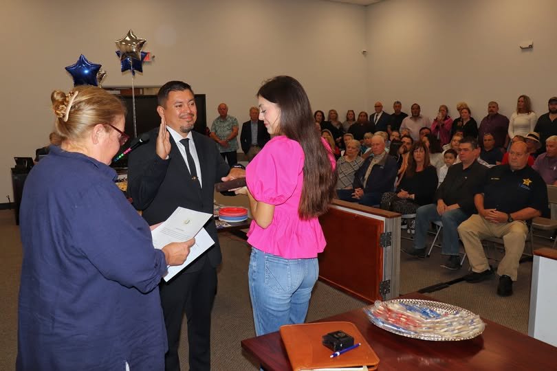 A woman in a blue shirt is reading from a paper, a man in a black suit with a badge is raising his right hand, and a woman with long hair in a pink top is receiving an oath or certificate in a room filled with seated and standing people. The room has