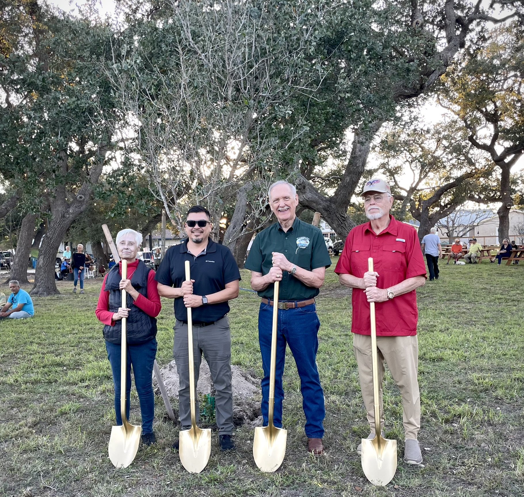 Four people standing outdoors in front of trees holding gold-colored shovels, smiling for a photo, with a group of people sitting and standing in the background.