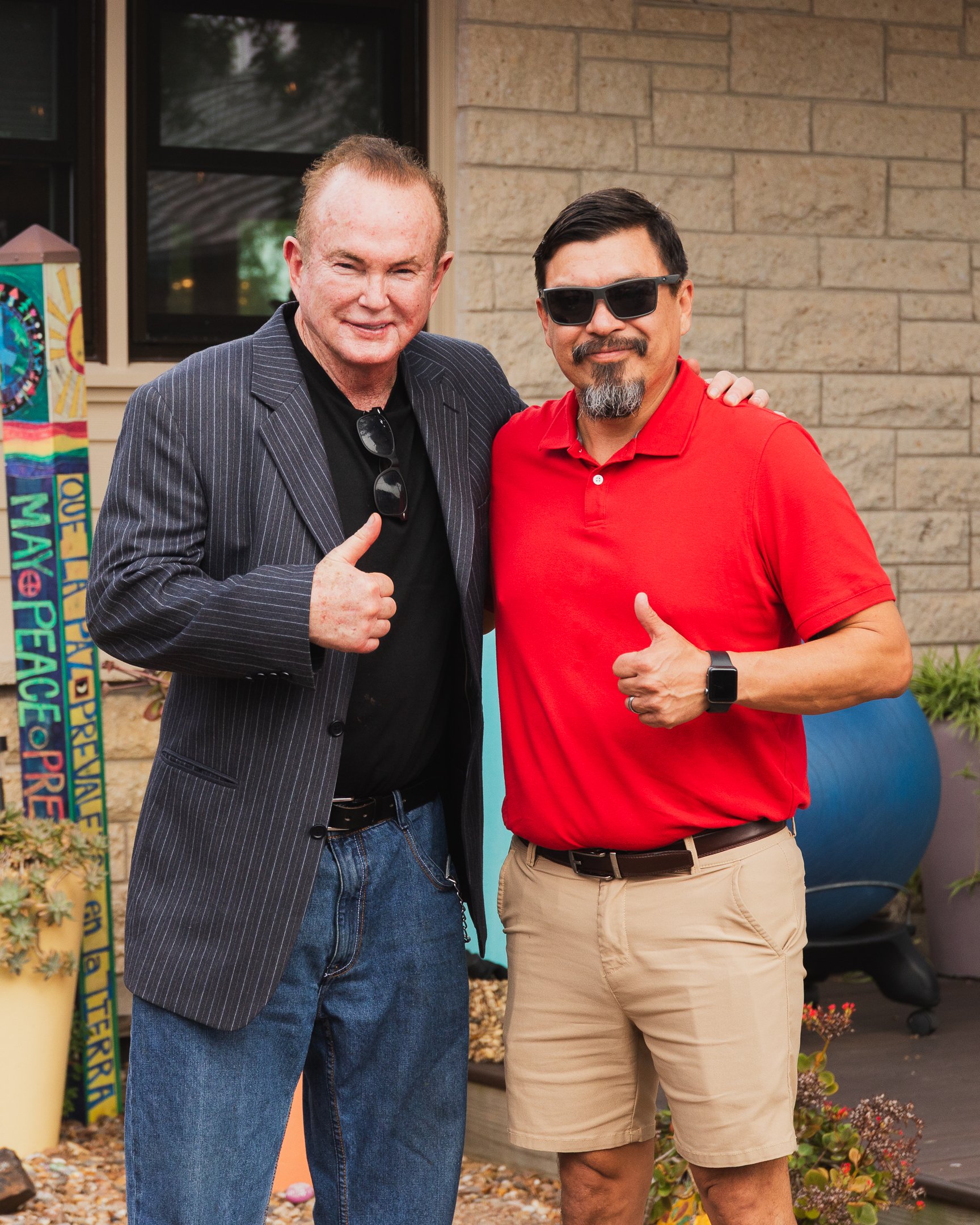 Two men standing outdoors in front of a tan brick building, giving thumbs up and smiling, one in a black pinstripe blazer and sunglasses, the other in a red polo shirt and sunglasses, with potted plants and a colorful sign nearby.