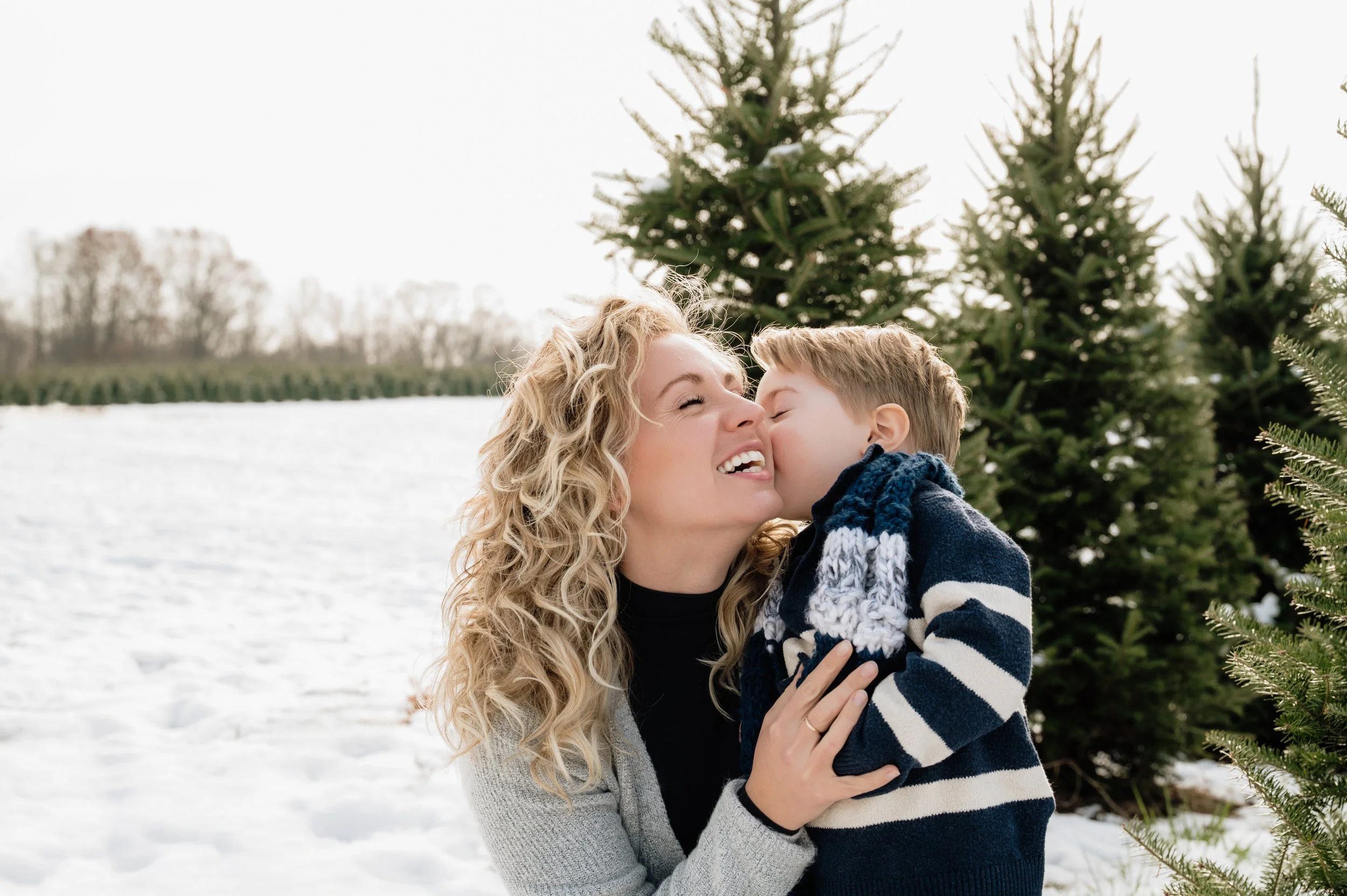 A woman with curly blonde hair smiling as a young boy kisses her on the cheek in a snowy outdoor setting with Christmas trees in the background.
