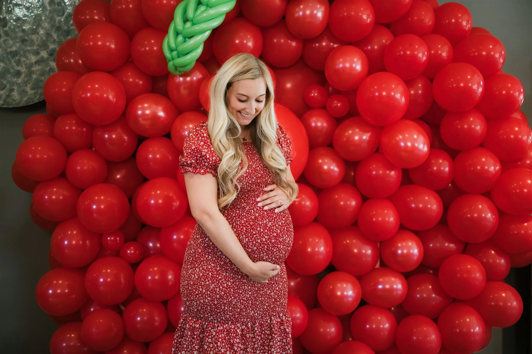 A woman in a red floral dress is smiling and holding her pregnant belly while standing in front of a wall decorated with numerous red and pink balloons and a large green balloon bow.