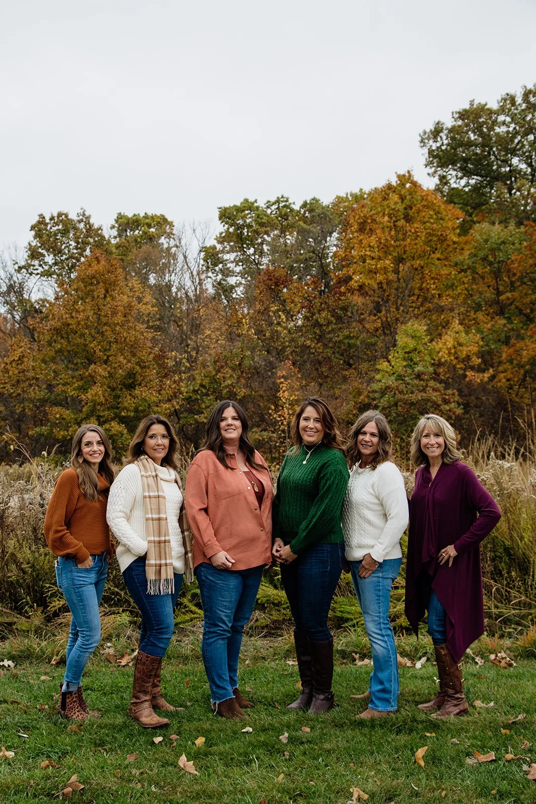 Six women standing outdoors in fall clothing on a grassy area, with trees showing autumn colors in the background.