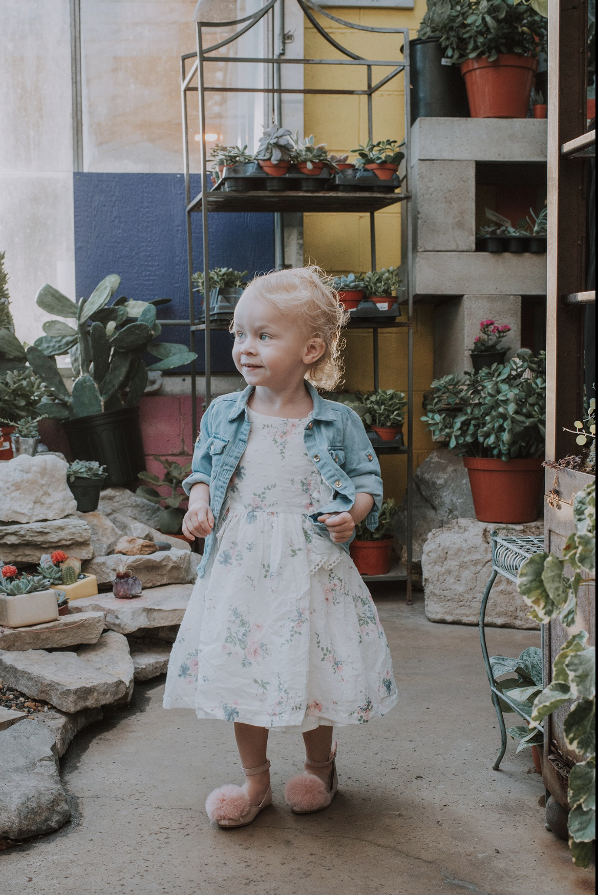 A young girl in a white dress with floral patterns, pink fuzzy shoes, and a denim jacket standing inside a plant nursery or greenhouse surrounded by potted plants and rocks.