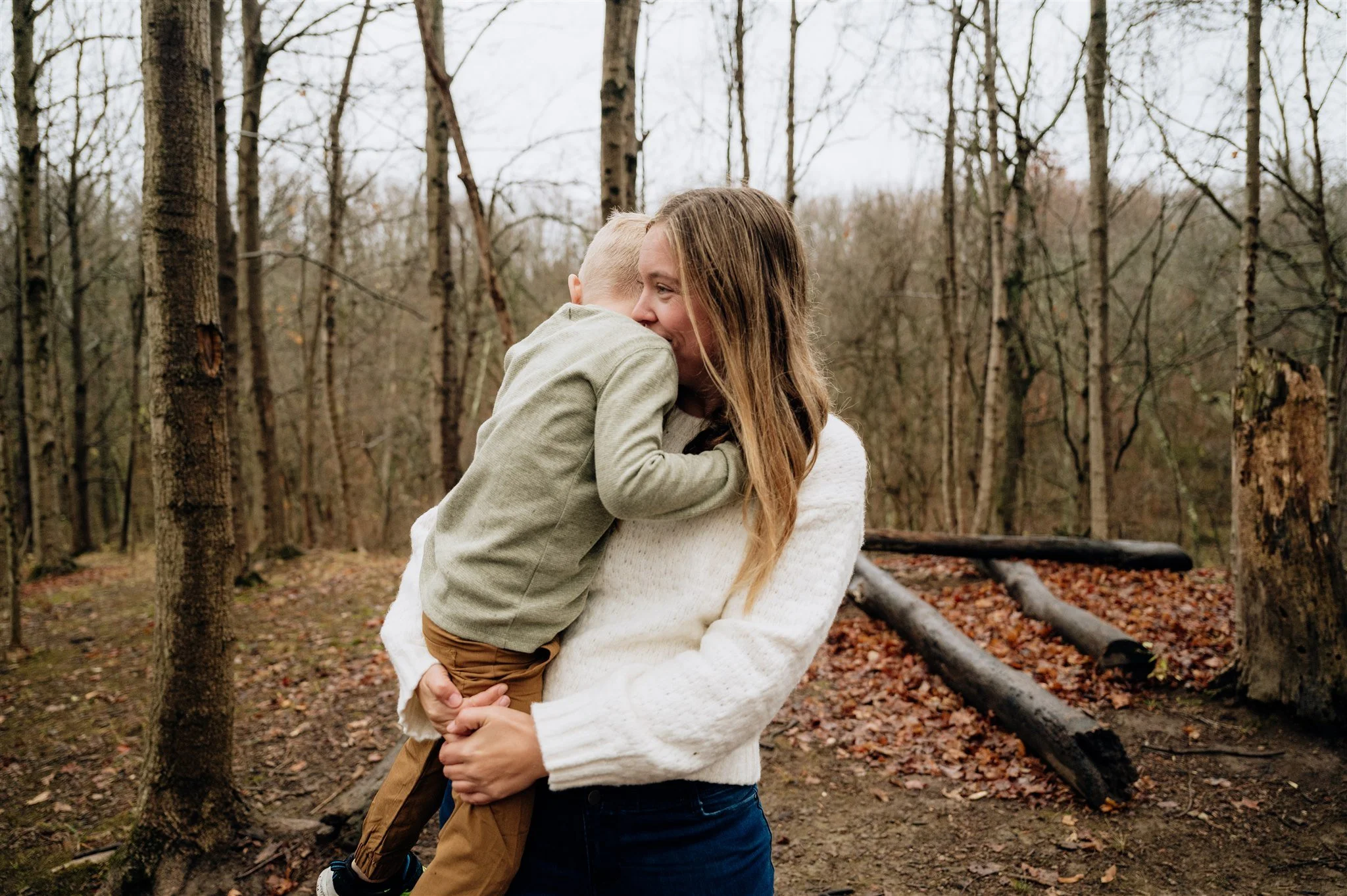 A woman holding a young boy in a forest, sharing a hug and smiling. The scene is autumn with fallen leaves and trees around.