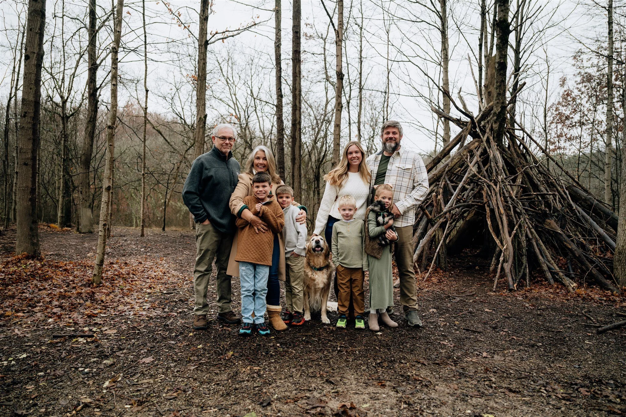 Family of eight with four children and two dogs standing in a forest in front of a teepee made of sticks.