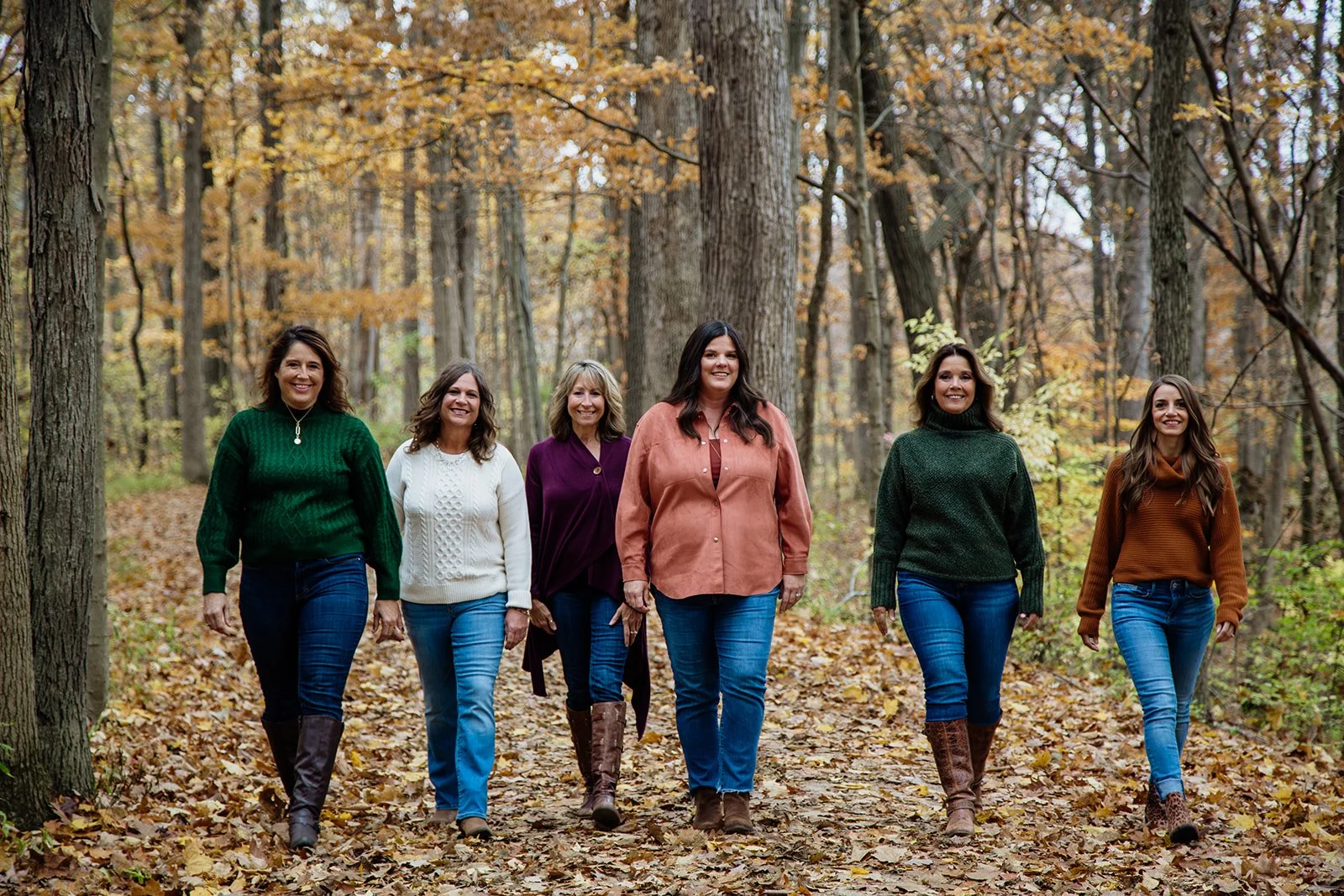 Six women walking on a leaf-covered trail through a forest in autumn, dressed in colorful sweaters and jeans, smiling and enjoying the fall scenery.
