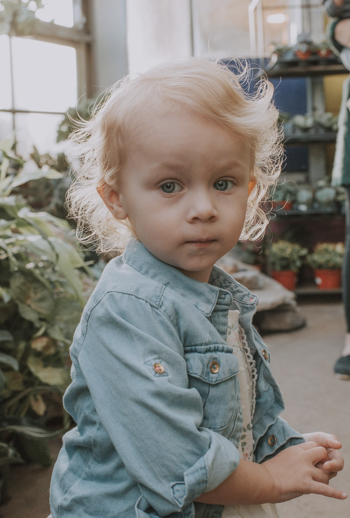 Young blonde girl with curly hair wearing a denim jacket in a greenhouse surrounded by various plants.