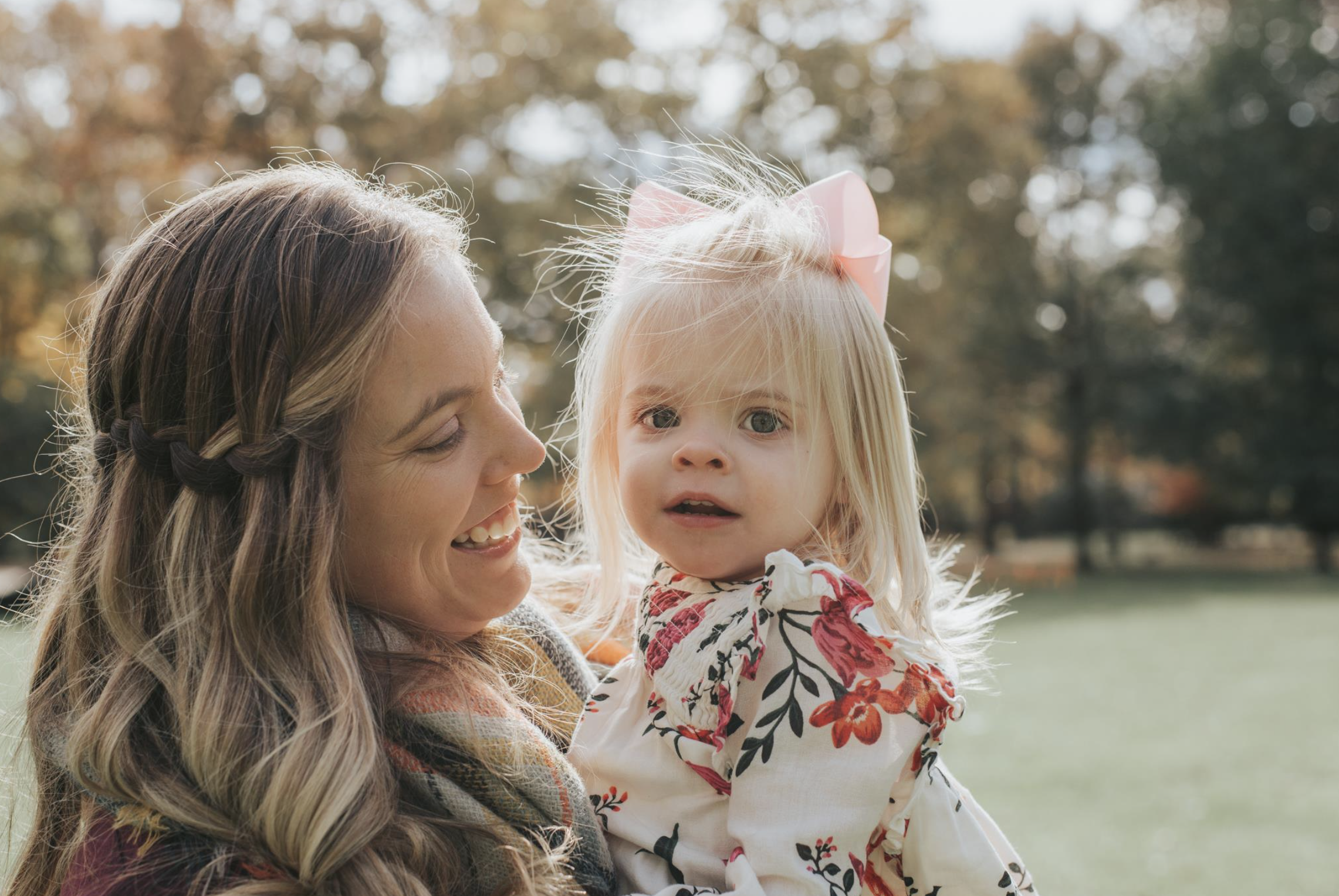Smile of woman holding a young girl with blonde hair and a pink bow in her hair, outdoors in a park with trees in the background.