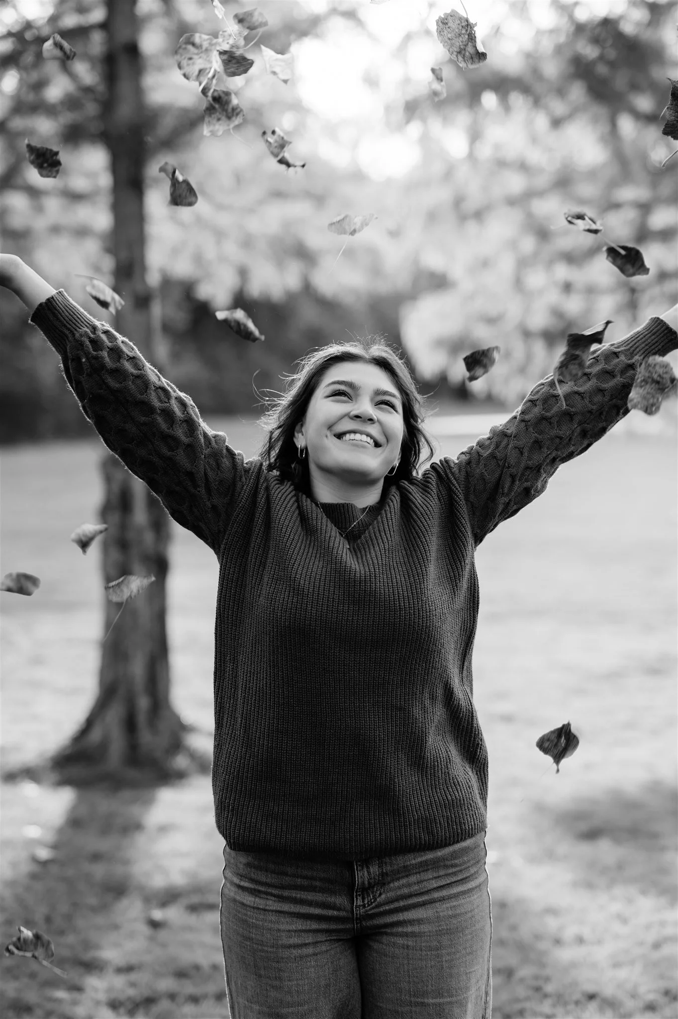 A woman outdoors smiling with her arms raised, surrounded by falling leaves.