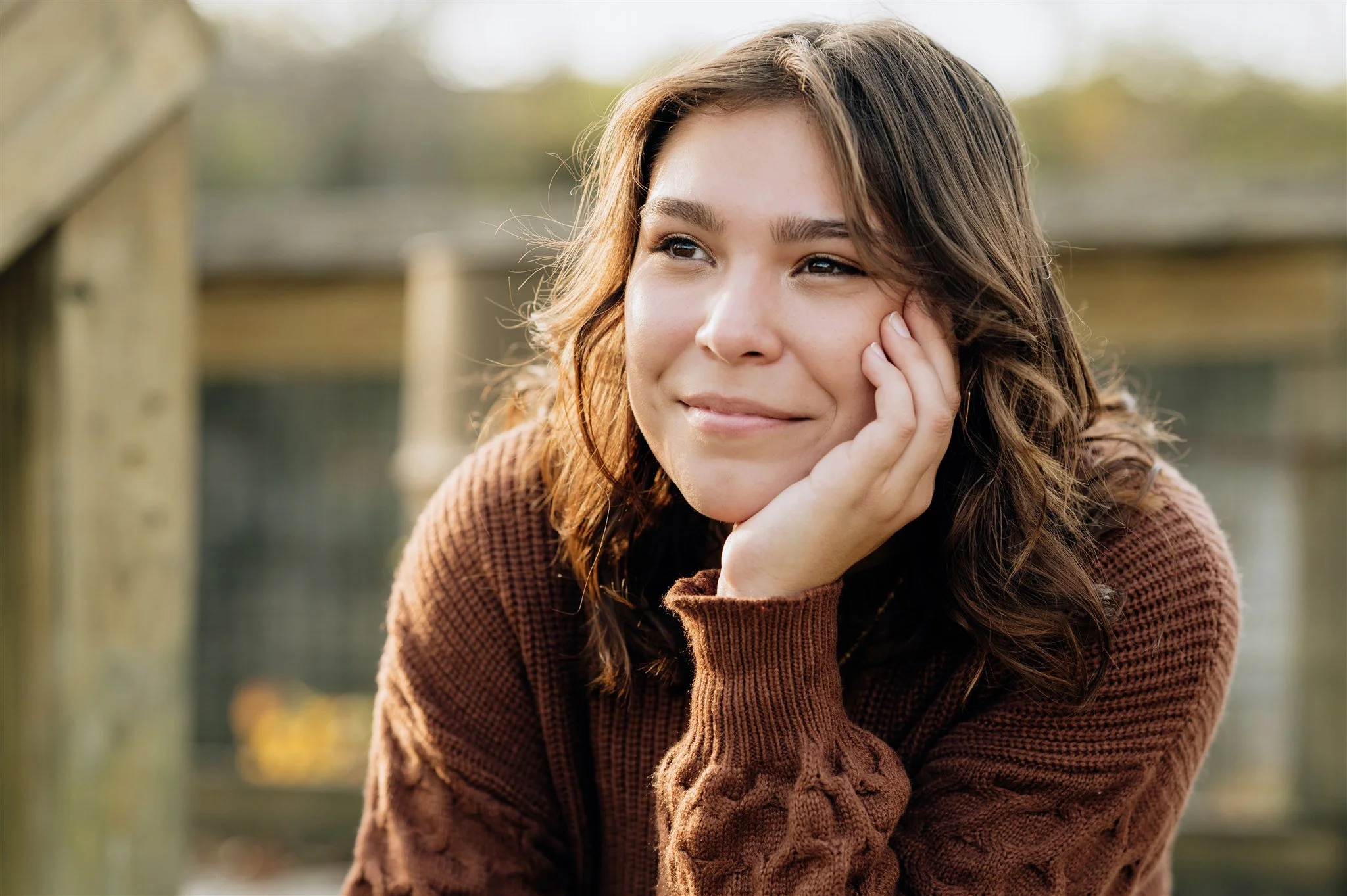 A woman with shoulder-length brown hair smiling with her head resting on her hand outdoors.