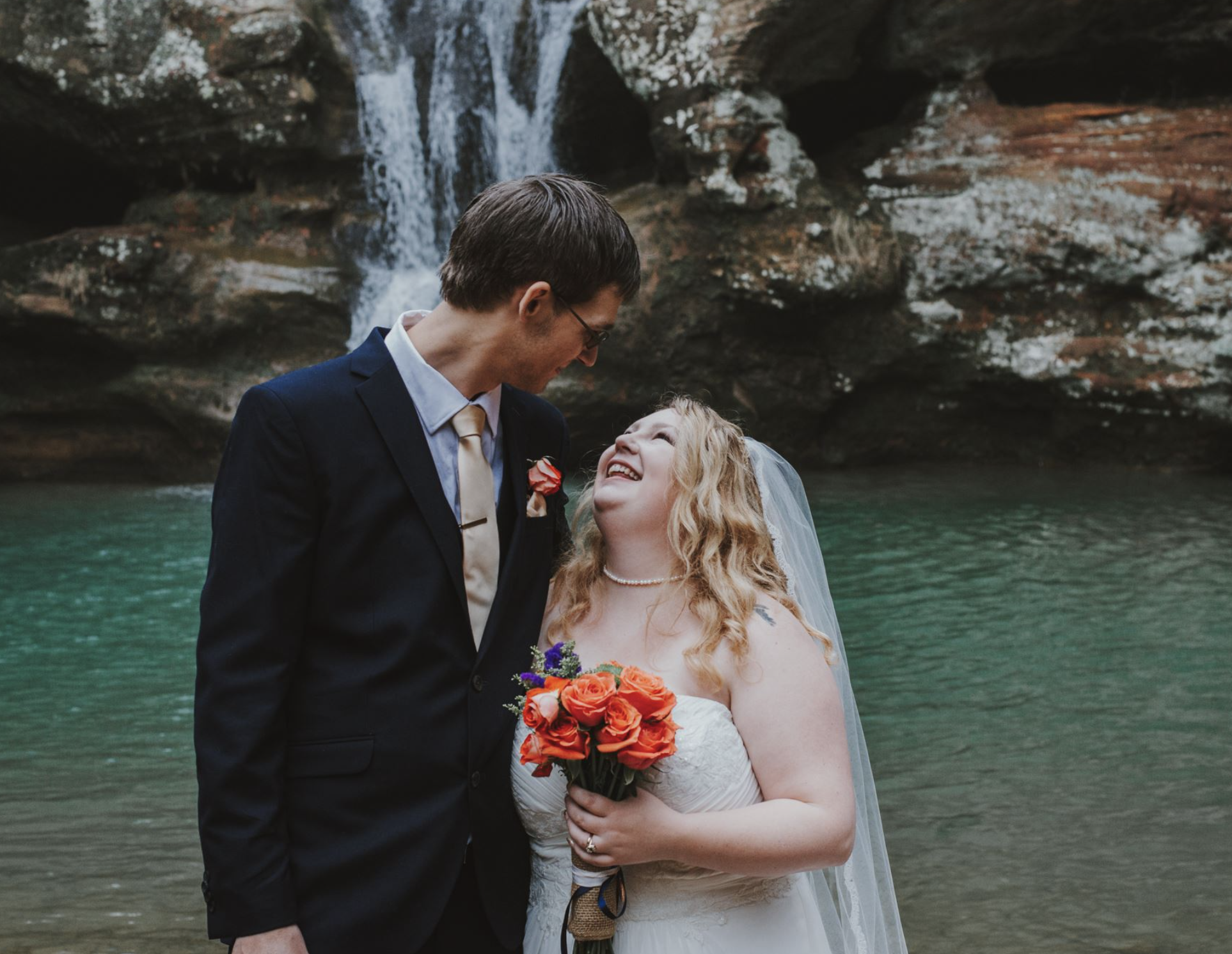 A bride and groom smiling and looking at each other by a waterfall and green water during their wedding.