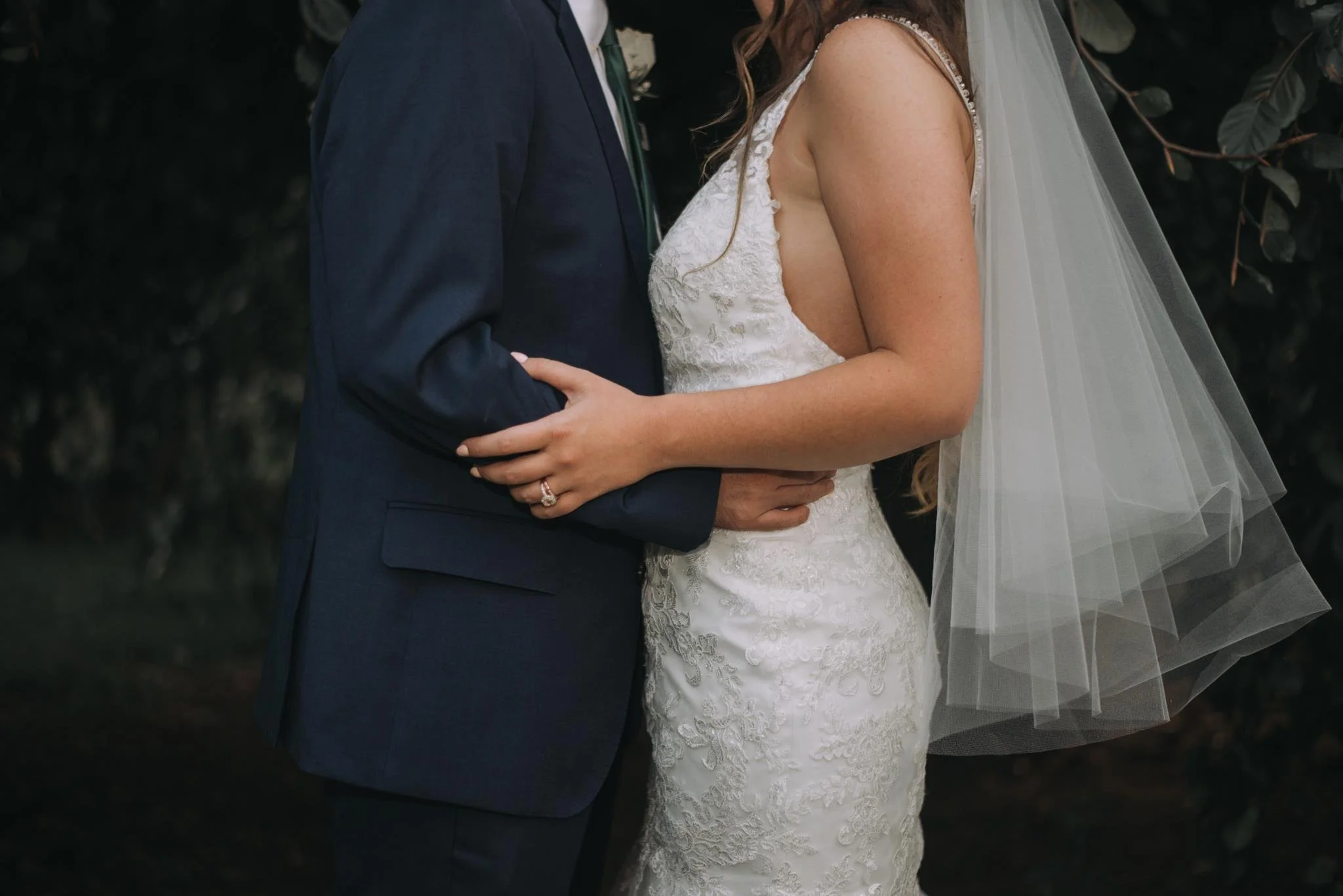 A bride and groom holding each other during their wedding ceremony, with the bride wearing a lace wedding dress and veil, and the groom in a navy suit. The couple's faces are not shown.