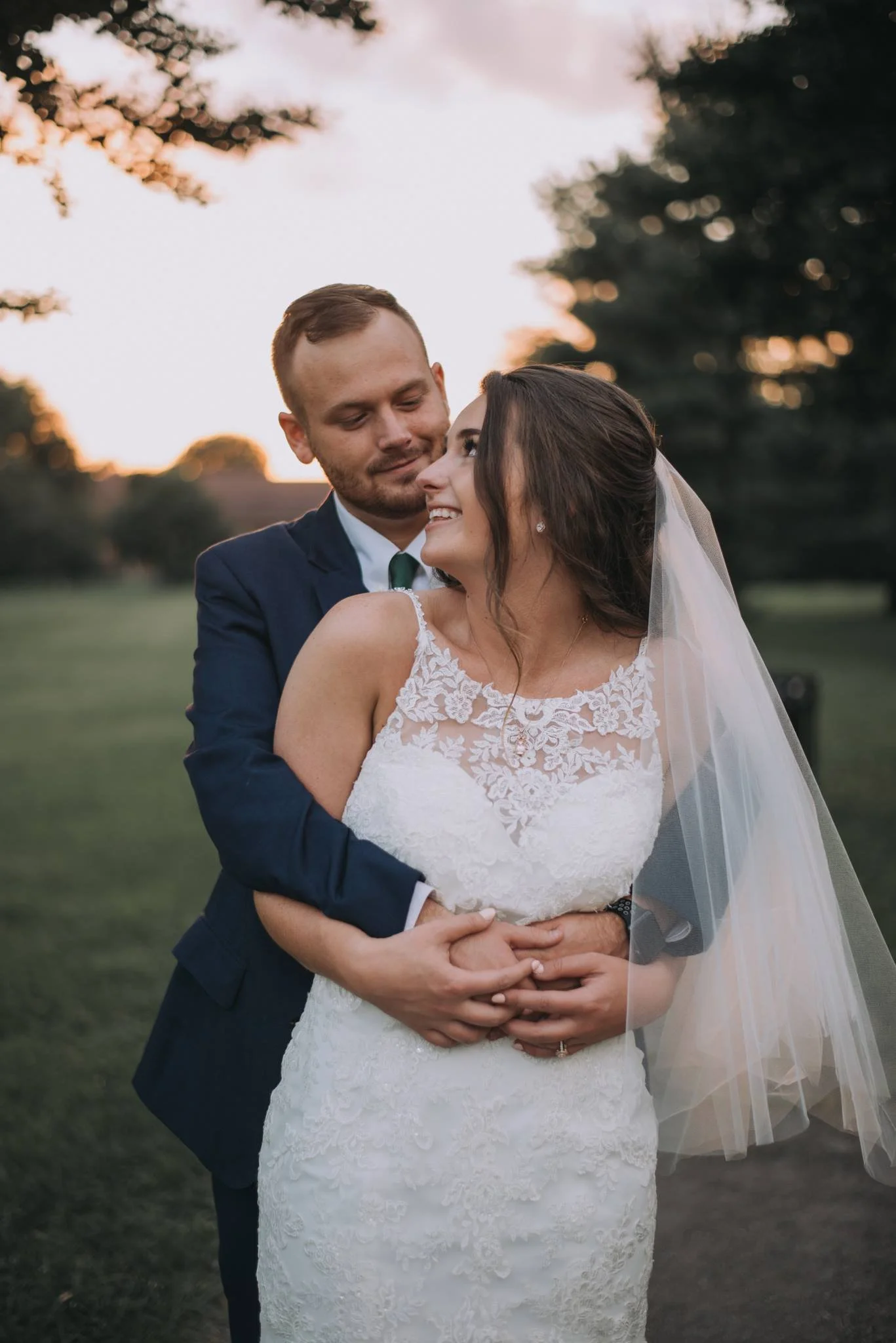 A bride and groom smiling and embracing outdoors during sunset, the bride wearing a white lace wedding dress and veil, the groom in a dark suit and tie, with a background of trees and a sunset sky.