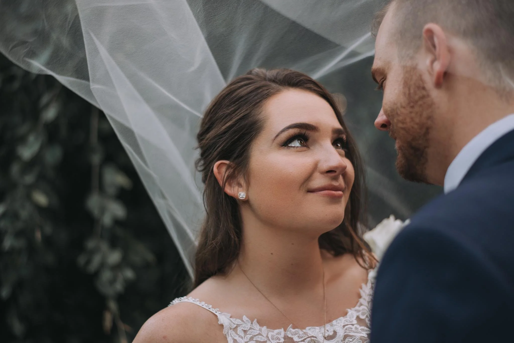 A bride and groom gazing at each other during their wedding ceremony. The bride has long brown hair, diamond earrings, and a white lace dress. The groom has short hair, a beard, and is in a navy suit. A white veil is visible in the background.