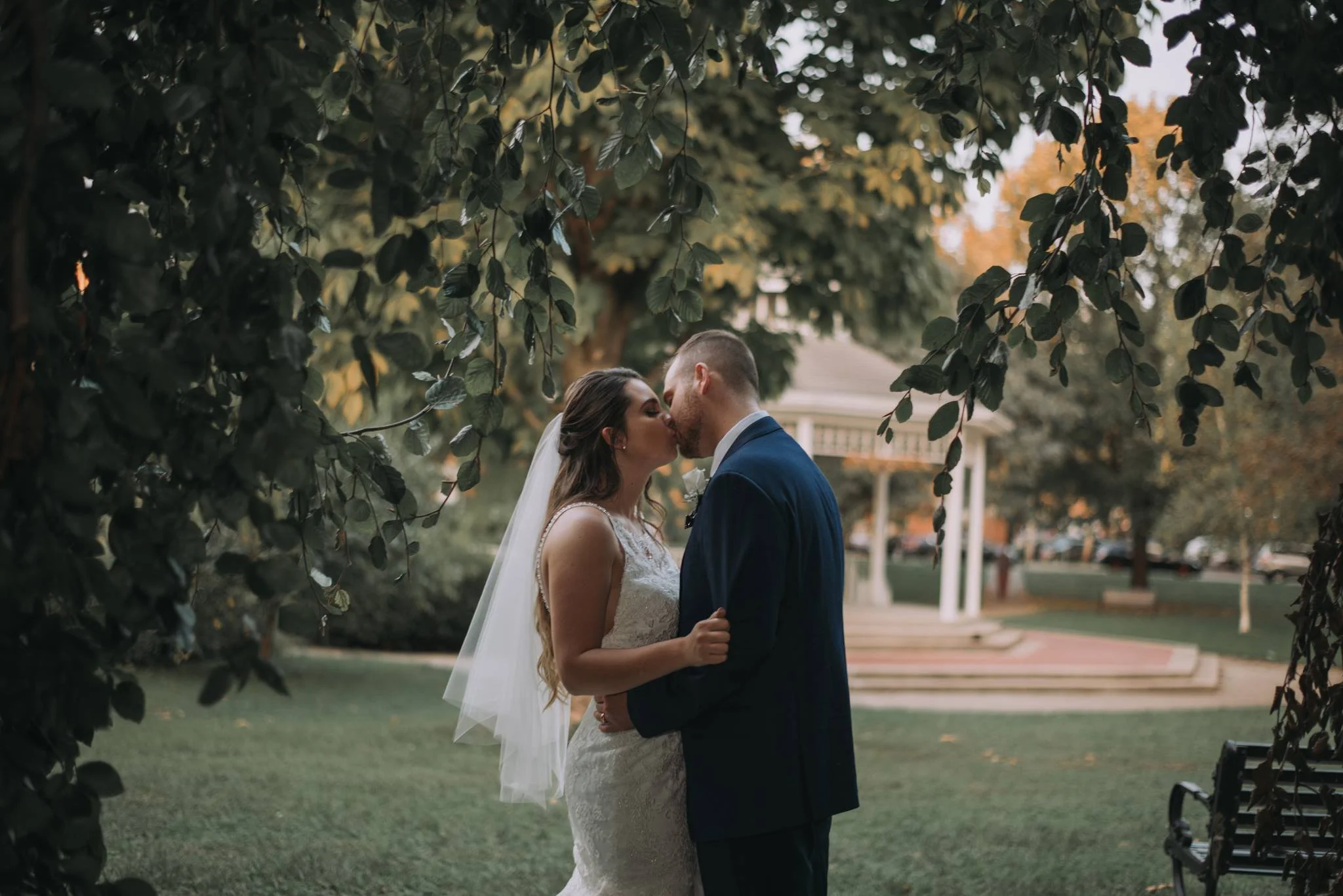 A bride and groom share a kiss under the trees in a park at sunset.