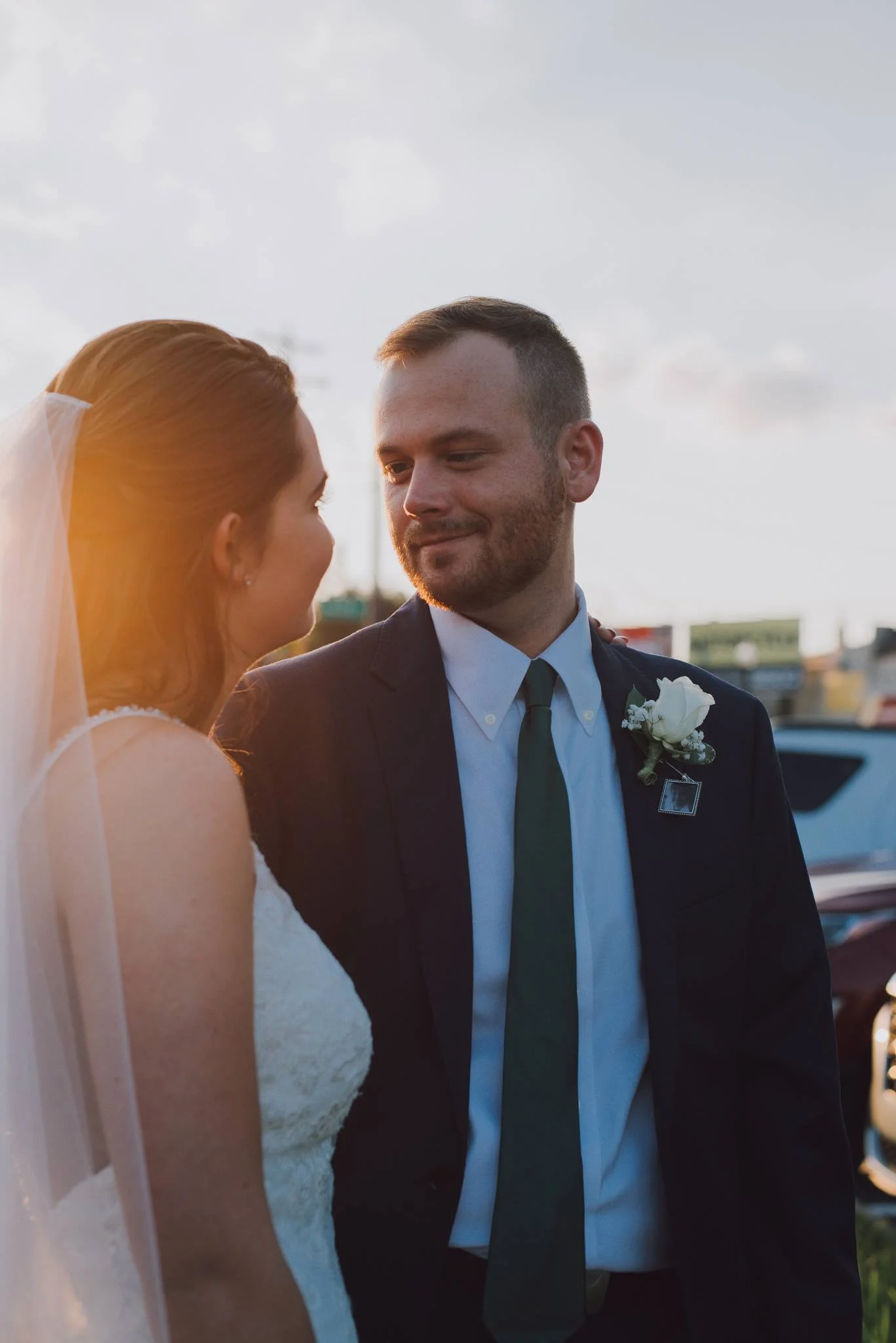 A bride and groom stand close together outside during sunset, gazing at each other with smiles, with cars and a partly cloudy sky in the background.