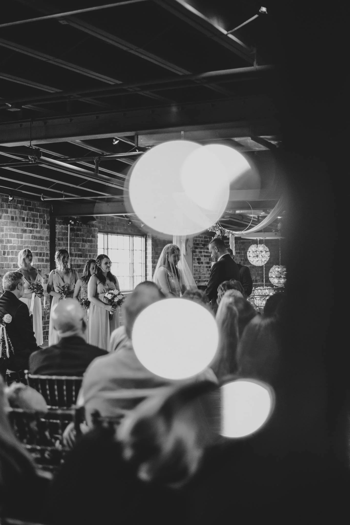Black and white photo of a wedding ceremony taking place indoors, with the bride and groom facing each other and holding hands, surrounded by bridesmaids and guests, with large circular lights in the foreground.