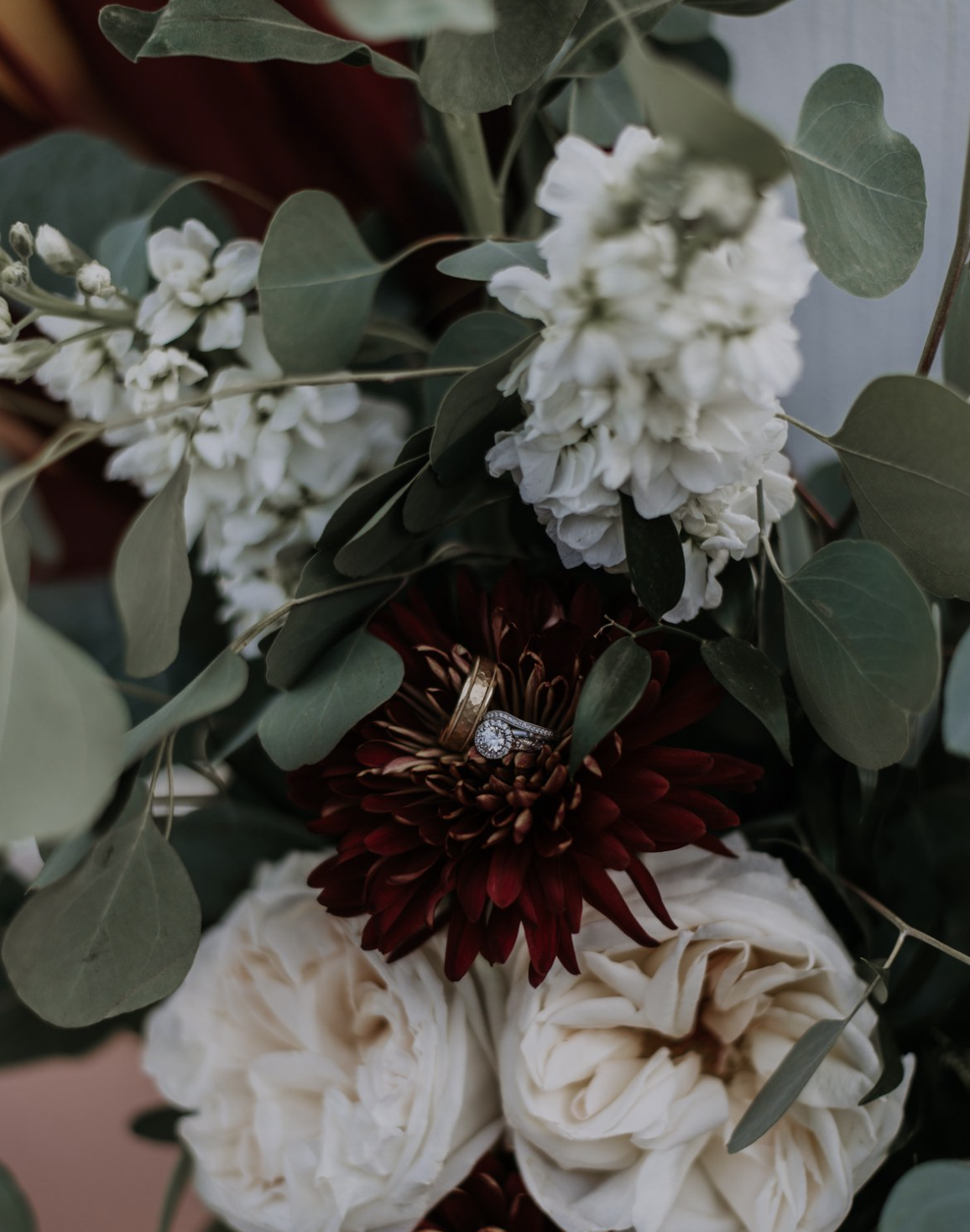 A bouquet of white, red, and cream flowers with green leaves, with a wedding ring placed on a dark red flower.