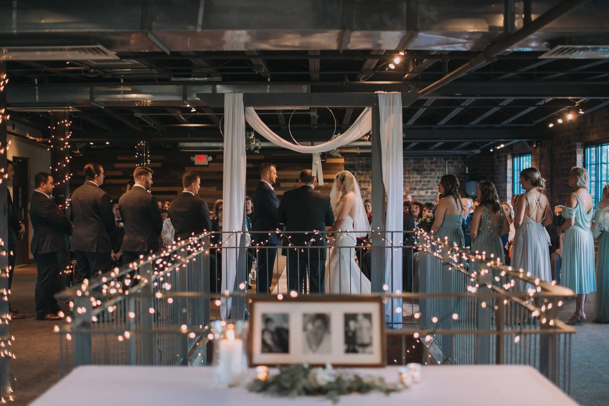 A wedding ceremony taking place in an indoor venue with a decorated altar and guests seated on either side. The bride and groom are standing in front of an officiant inside a canopy with white drapes, while bridesmaids and groomsmen stand nearby.