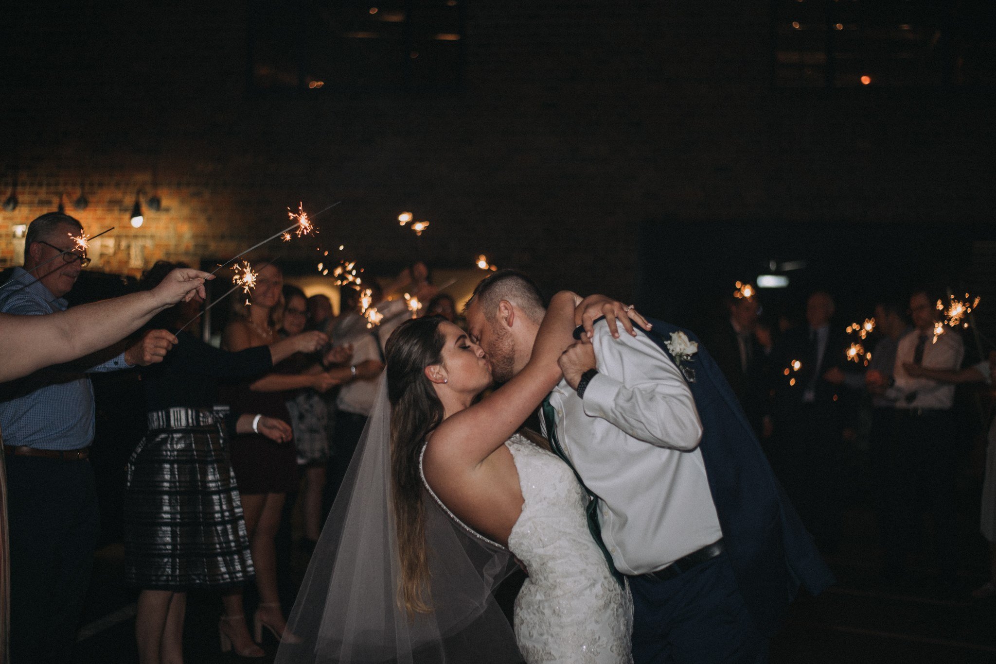 a bride and groom kissing at their wedding reception surrounded by guests holding sparklers in a dark indoor venue