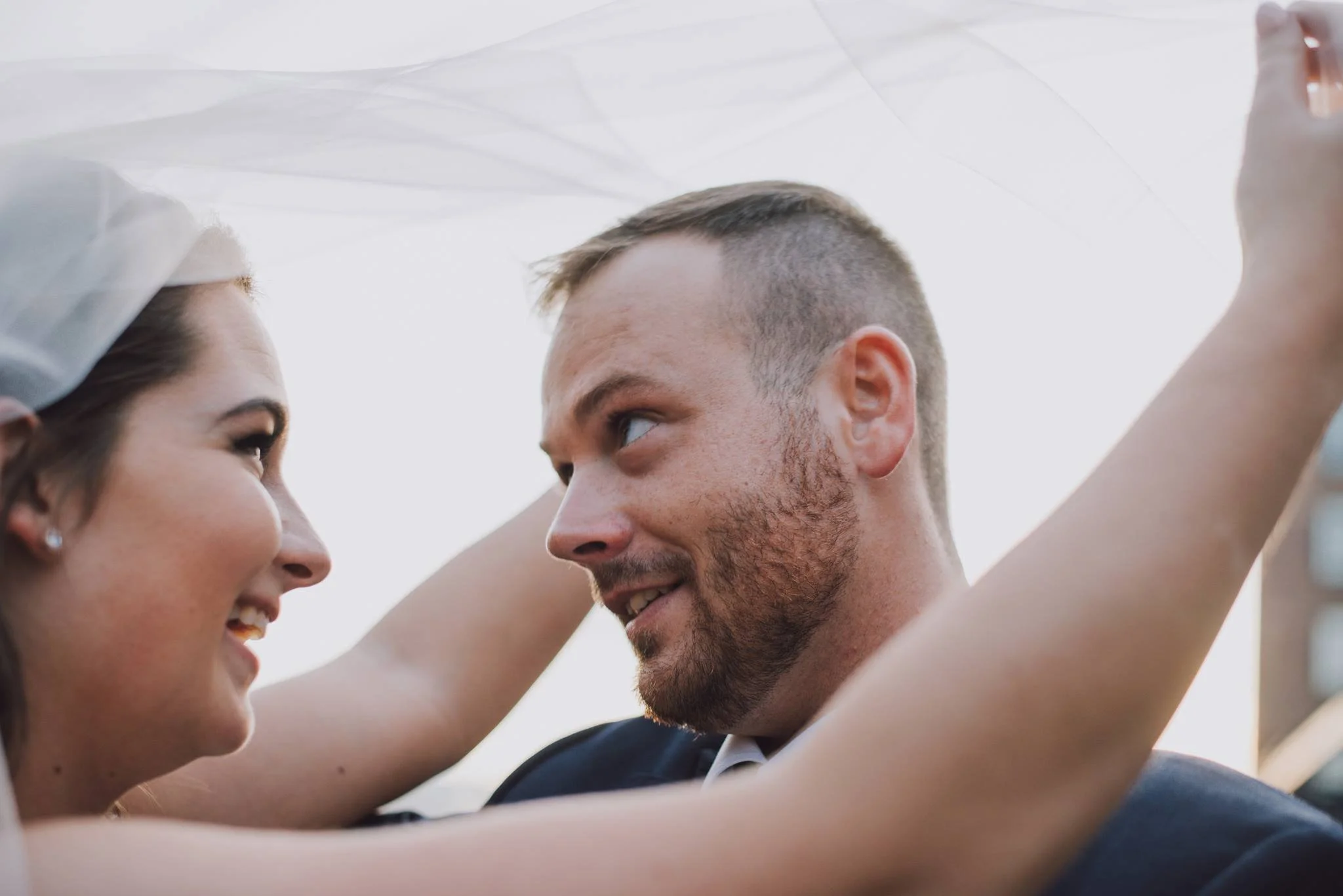 A bride and groom smiling at each other during their wedding, with the bride placing a veil over the groom's head.