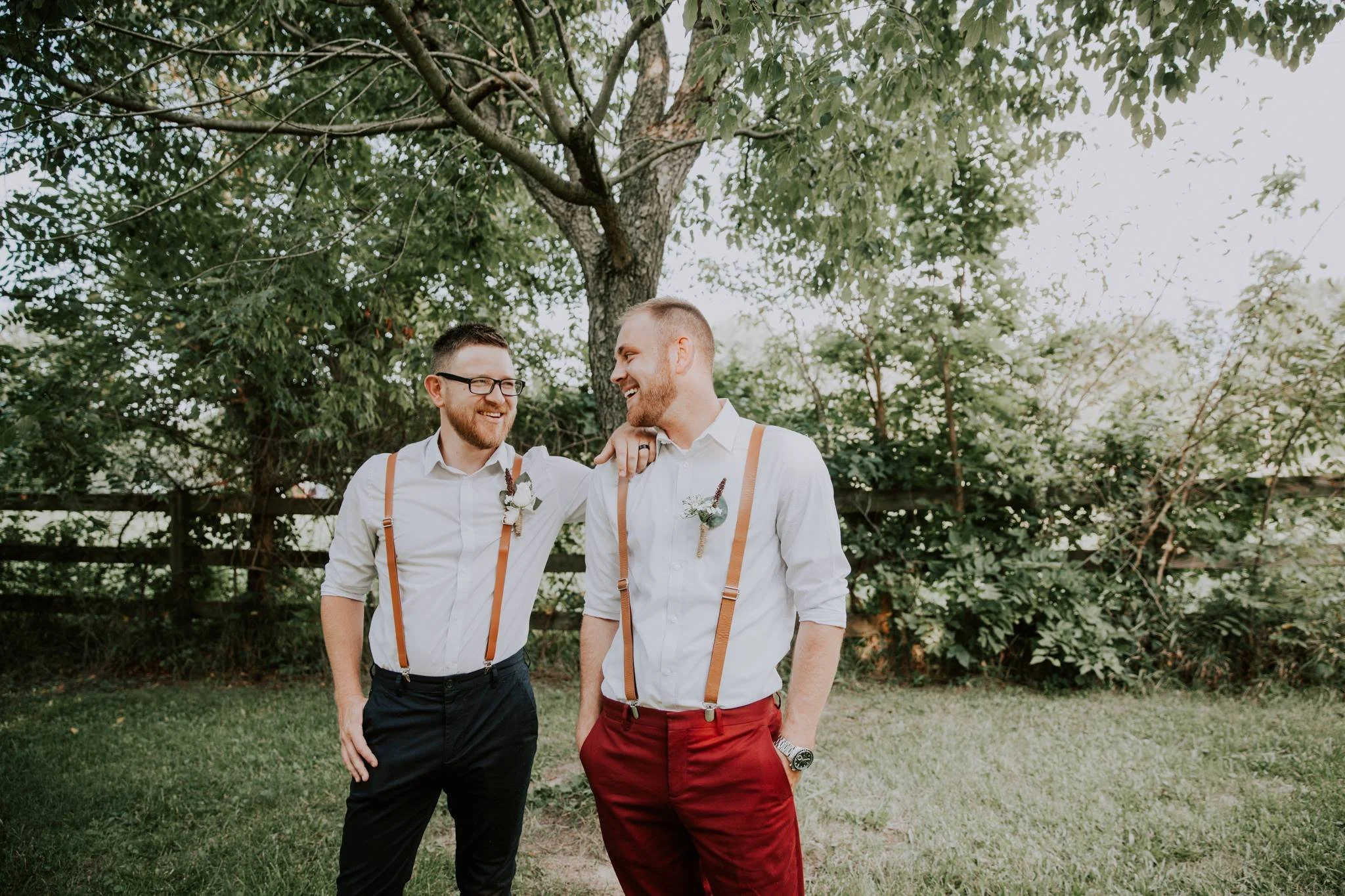 Two men in white shirts and suspenders, smiling and enjoying each other's company outdoors near a large tree and green bushes.