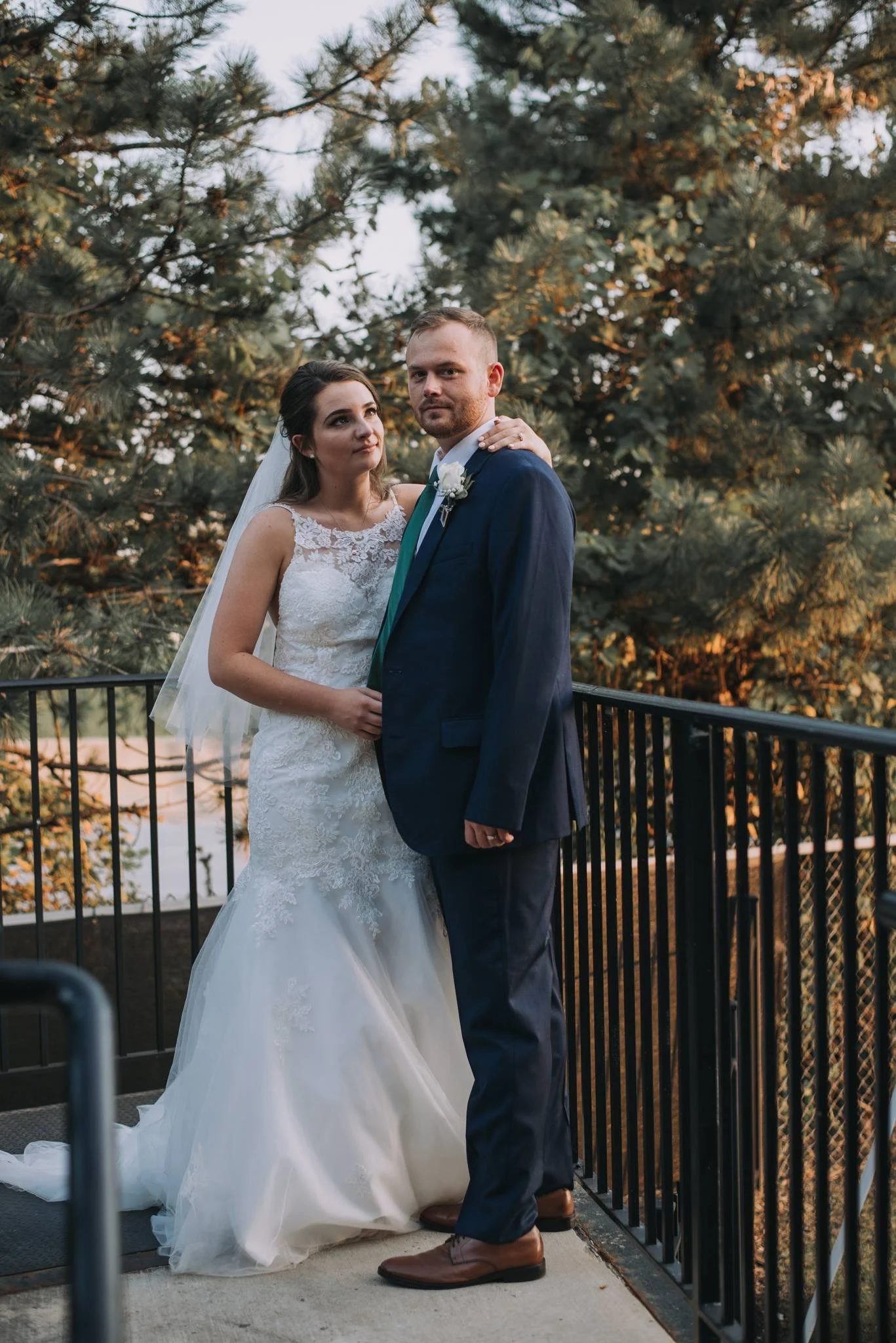 A bride and groom standing on a balcony outdoors at sunset, with trees behind them. The bride wears a white lace wedding dress with a veil, and the groom wears a blue suit with a white shirt and brown shoes.