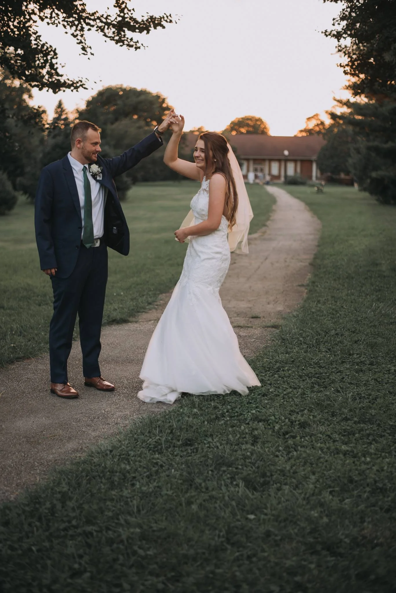 A bride and groom dancing outdoors during sunset on a pathway, with trees and a house in the background.