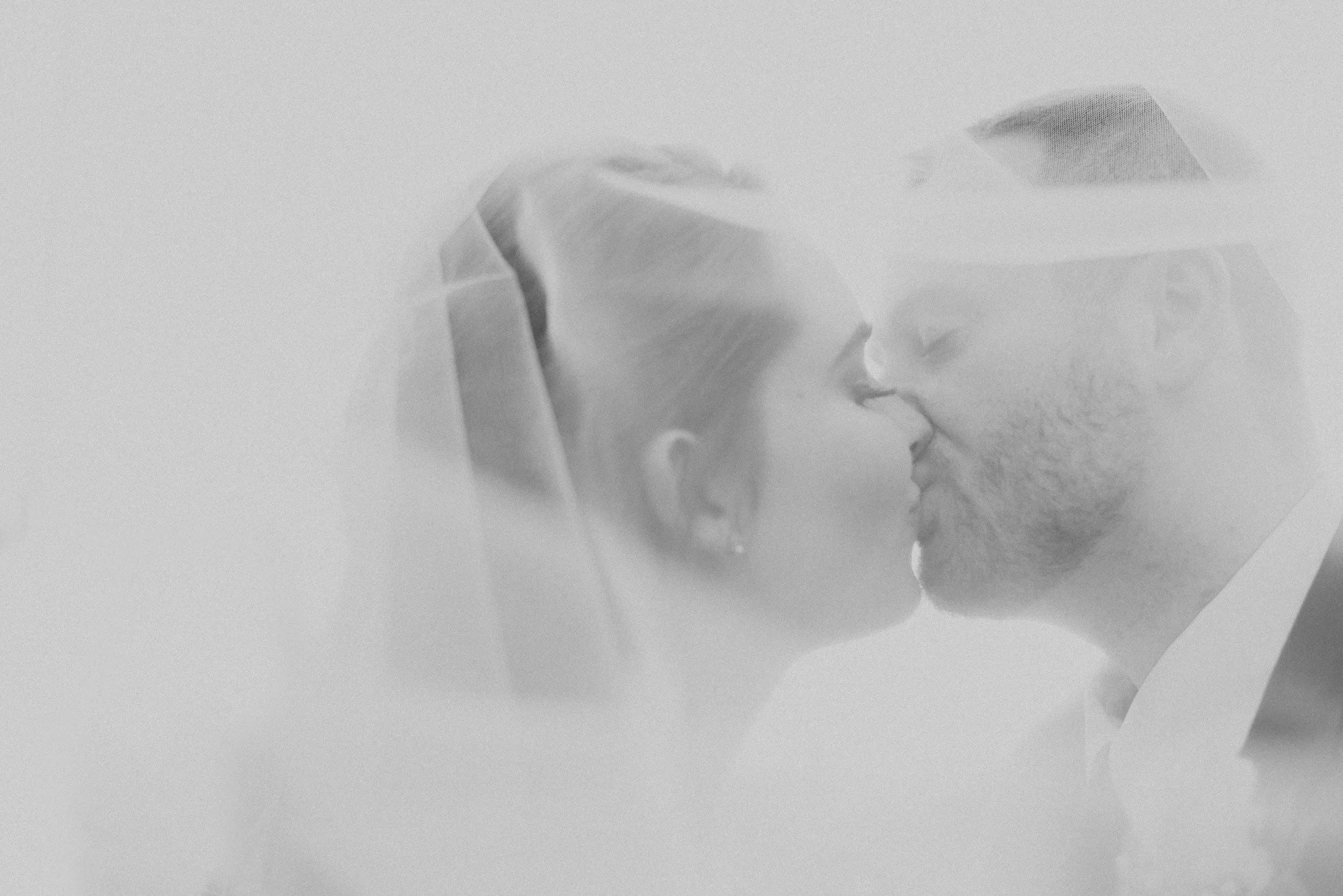 Black and white photo of a bride and groom about to kiss, with the bride wearing a veil and earrings, and the groom wearing glasses and a suit.
