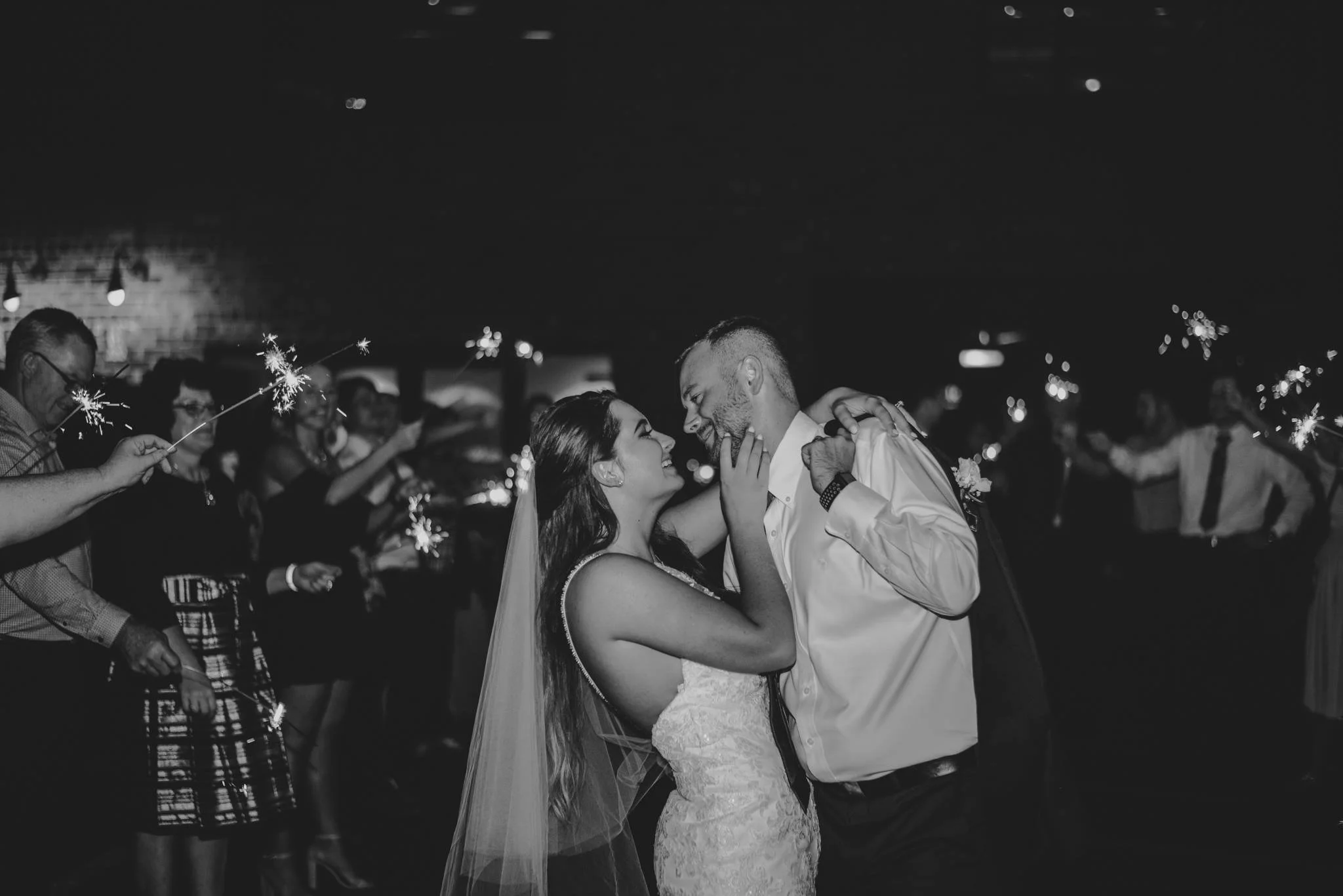 A bride and groom dancing and looking into each other's eyes at a wedding reception, with sparklers held by guests in the background.