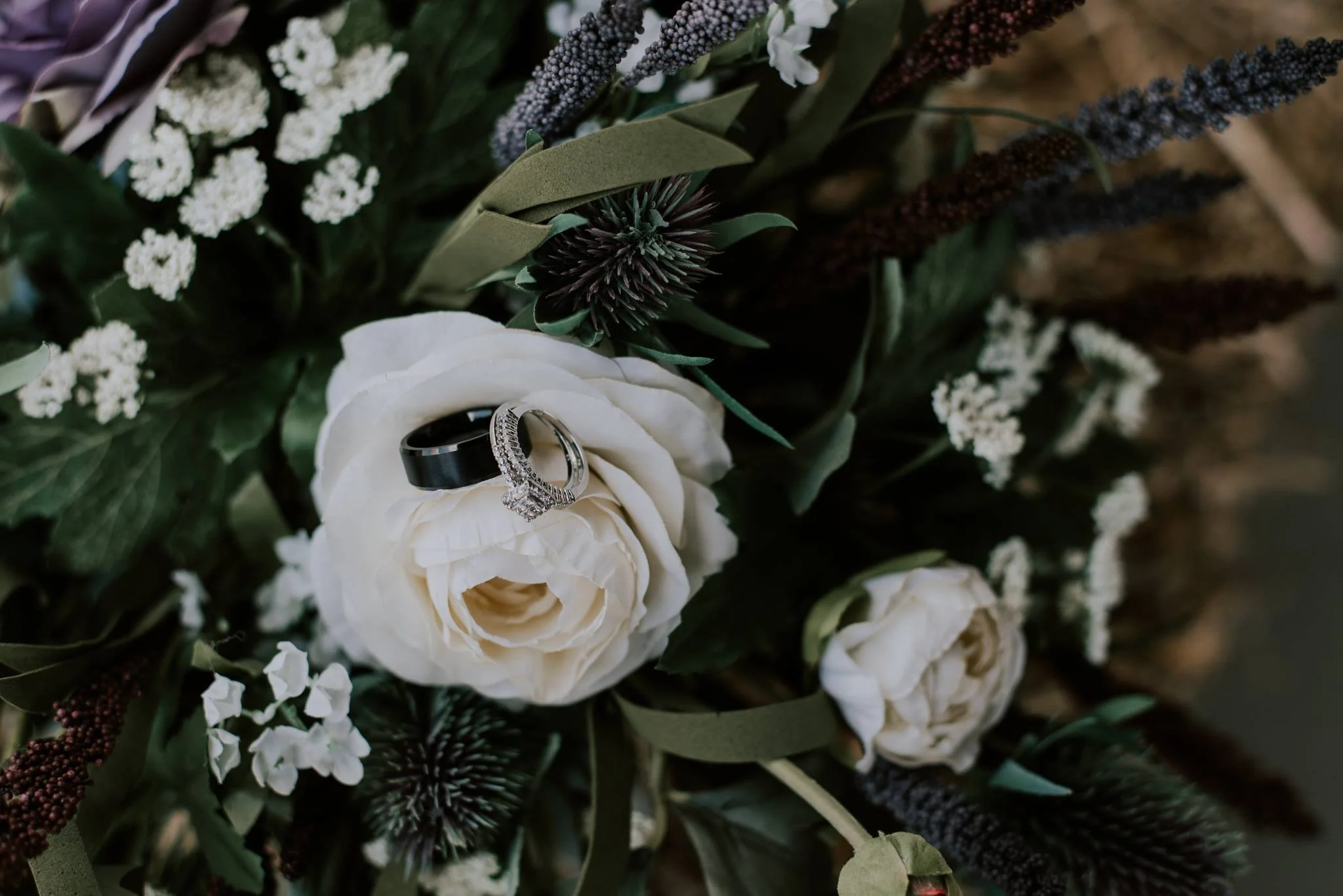A bouquet of white and purple flowers with two wedding rings, one silver with diamonds and one black, resting on a white rose in the bouquet.